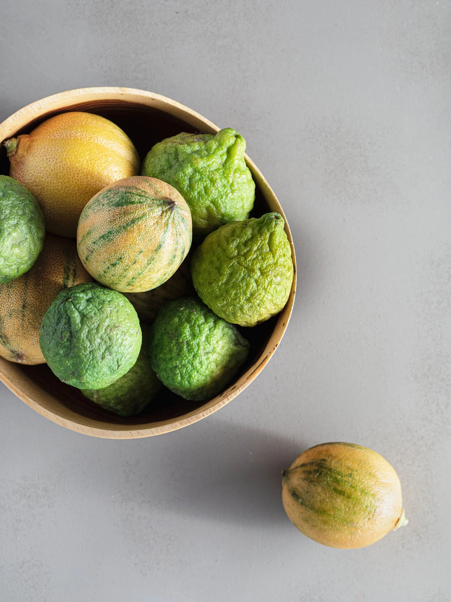 A wooden bowl filled with kaffir limes and calamansi limes, with one calamansi lime outside the bowl on a gray surface.