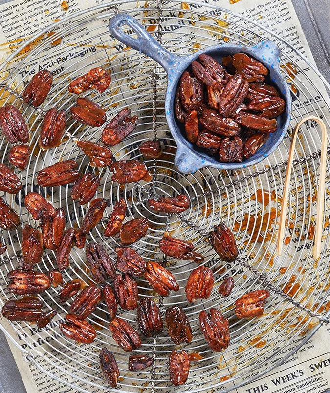 A wire cooling rack with roasted pecans, some in a small blue pot, and some spilled onto a newspaper.