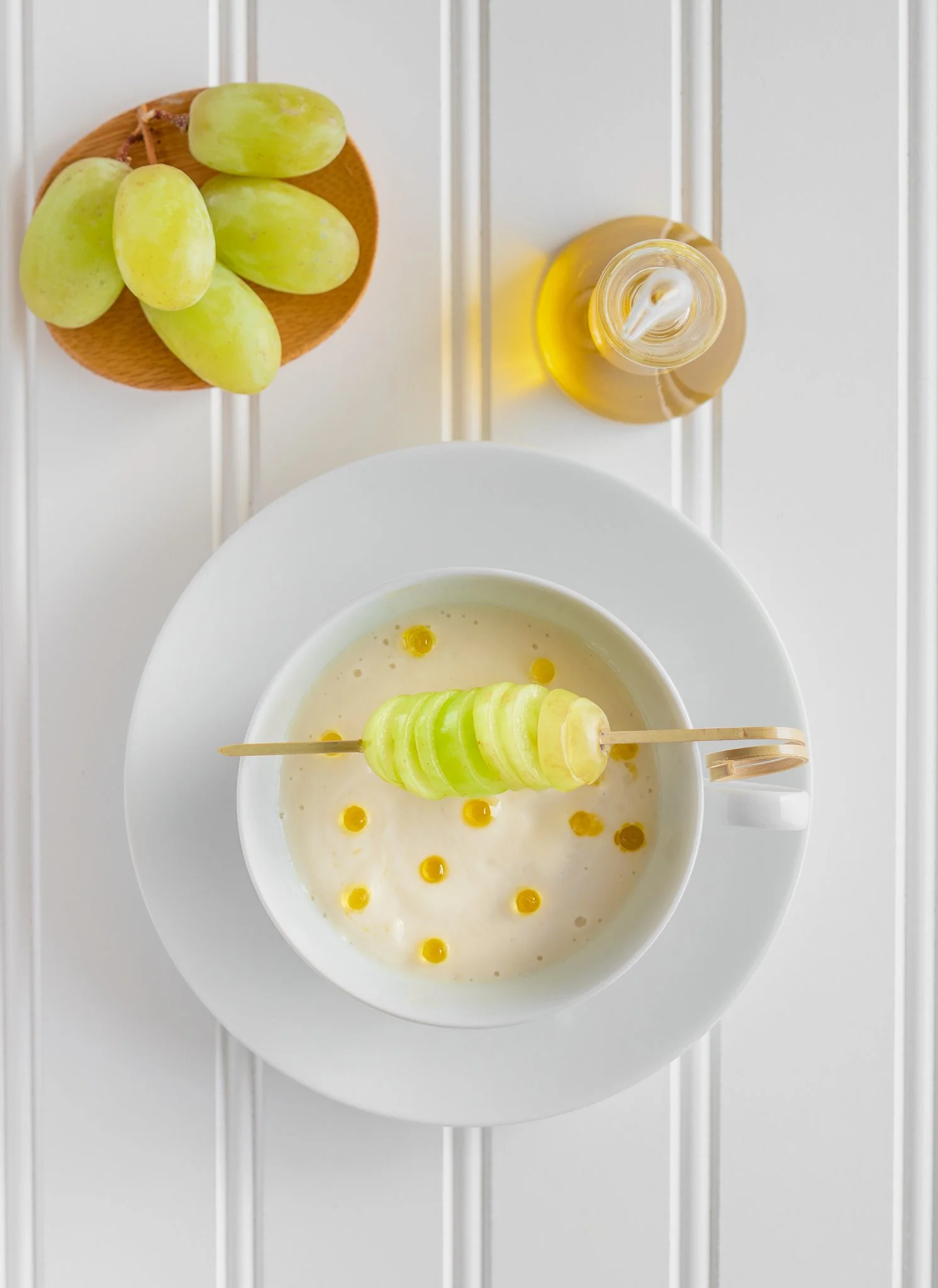 A bowl of sliced green grapes on a skewer in yogurt with yellow and orange toppings, with a small plate of additional green grapes and a bottle of yellow liquid next to it, all on a white surface.