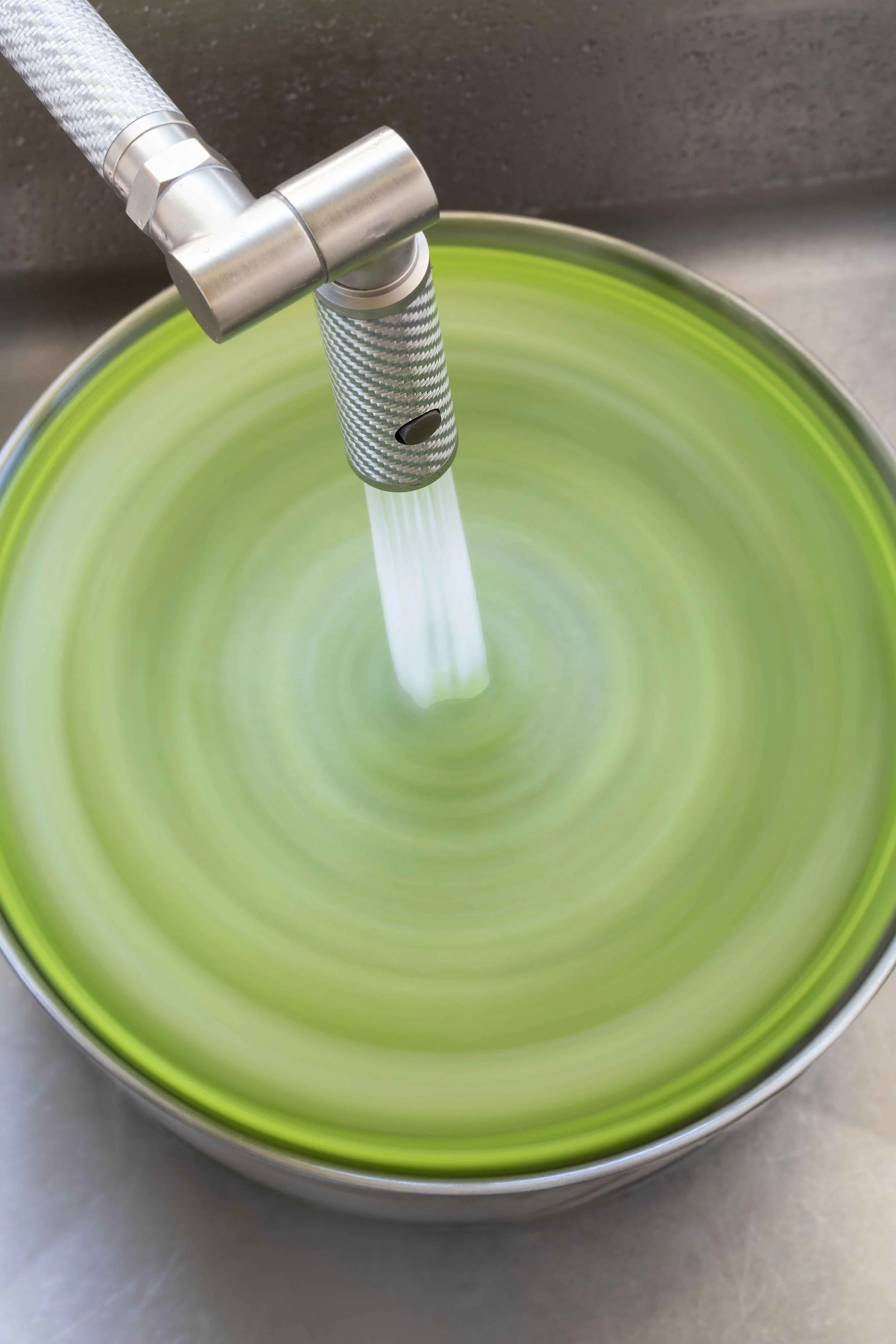 A close-up of a kitchen sink faucet with water flowing into a green basin, captured from above.
