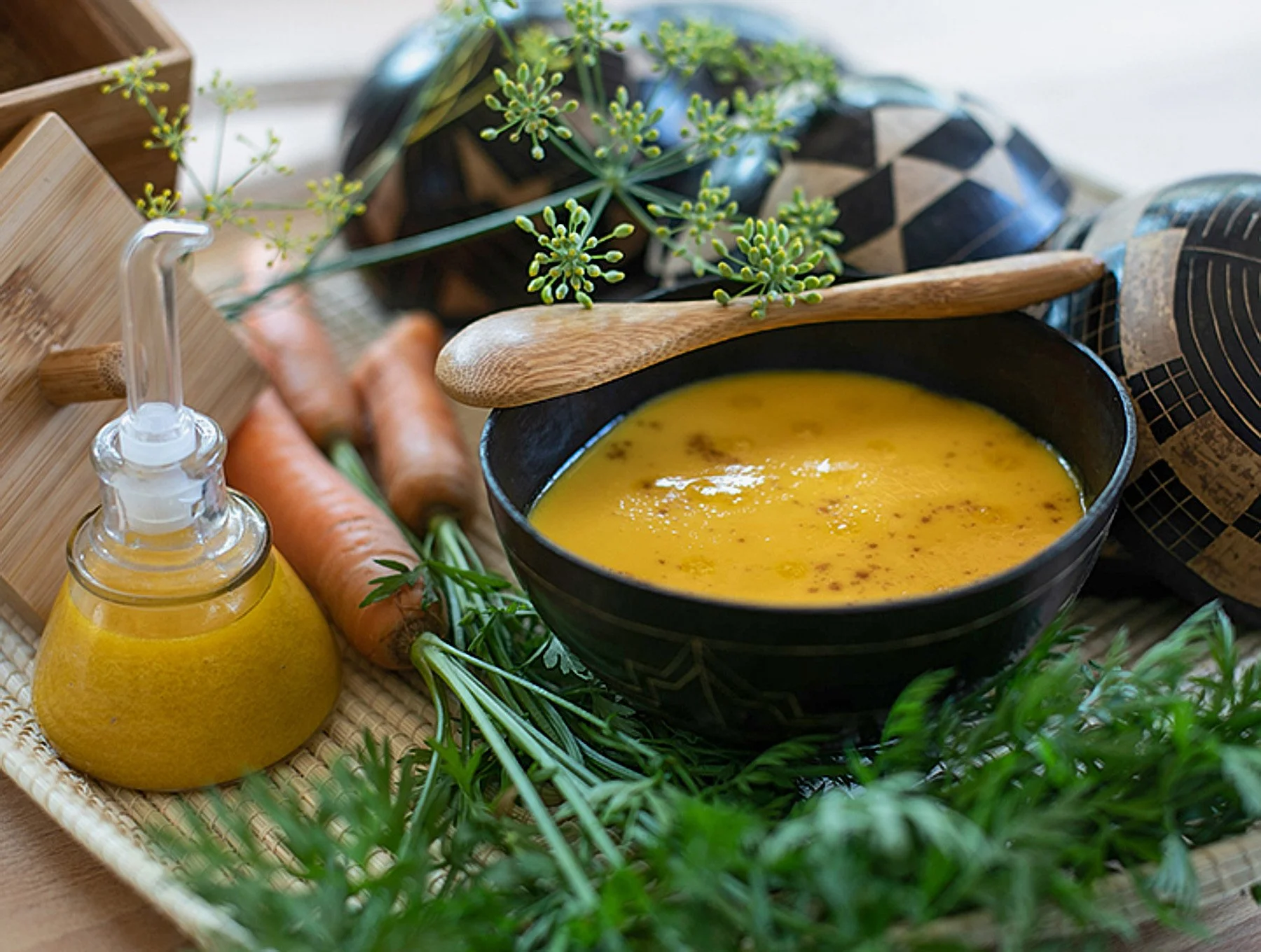 A black bowl of yellow soup, a wooden spoon resting on the bowl, surrounded by fresh carrots, greenery, a dropper bottle of yellow liquid, and a cloth with a geometric pattern, all placed on a woven tray.