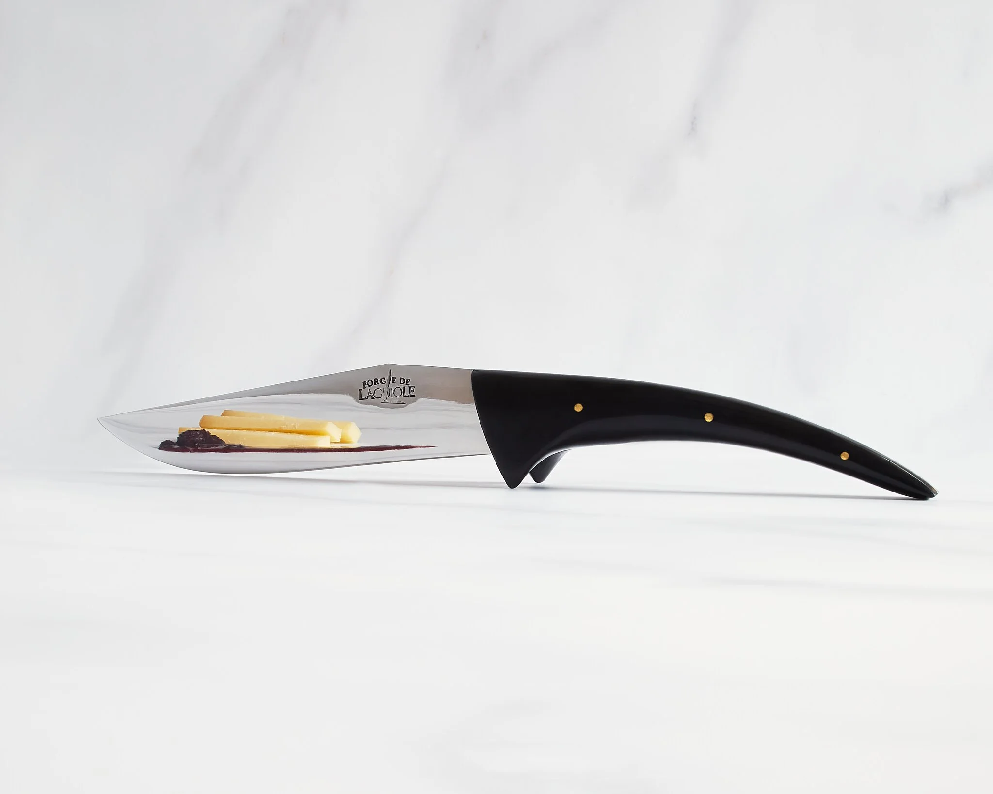 A knife with a black handle and a stainless steel blade, resting on a white surface against a white marble background. The blade has inscriptions that read "FORGE DE LACALLO" and appears to contain some butter and a dark jam or berry spread.