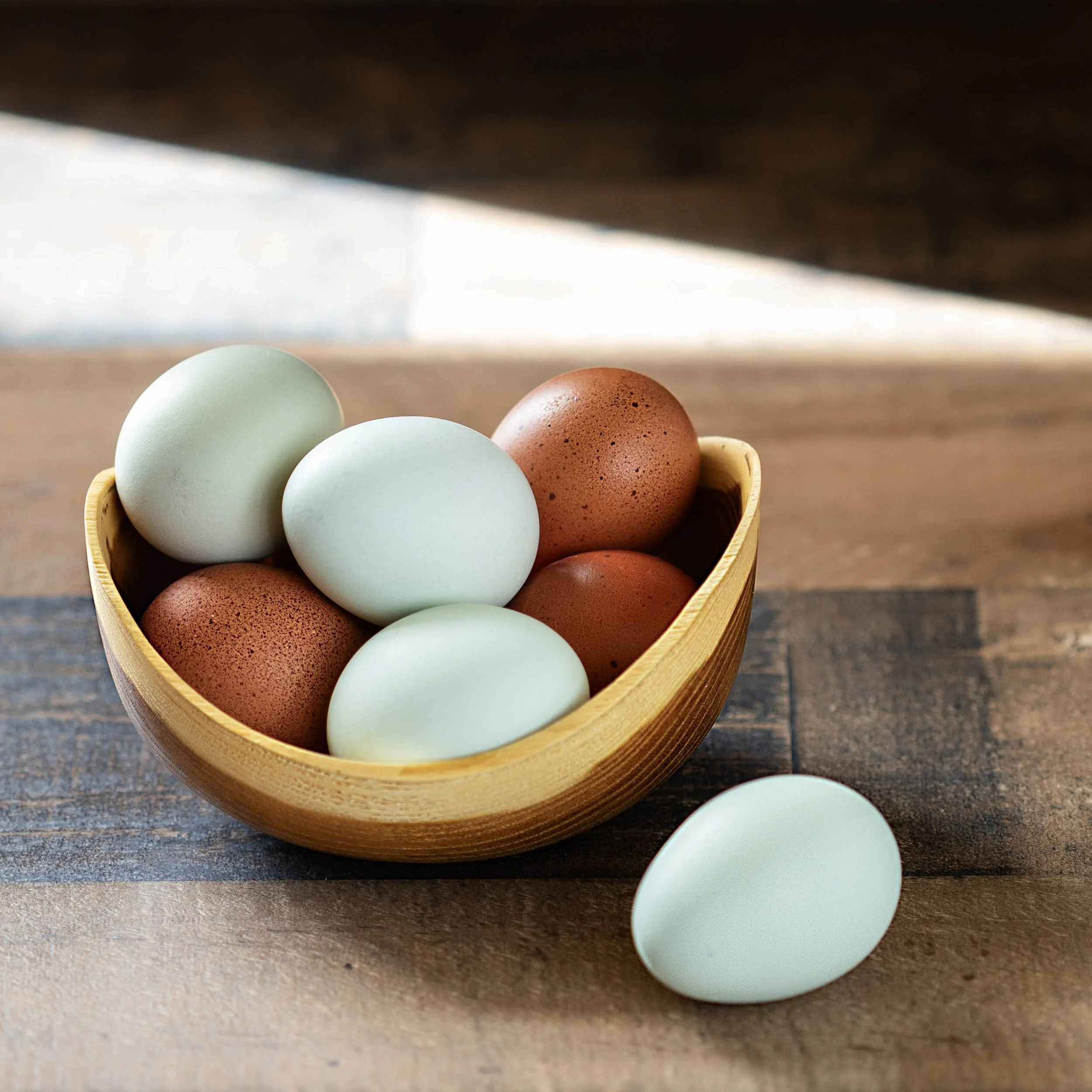 A wooden bowl filled with a mixture of brown and white eggs, with one white egg outside the bowl on a wooden surface.