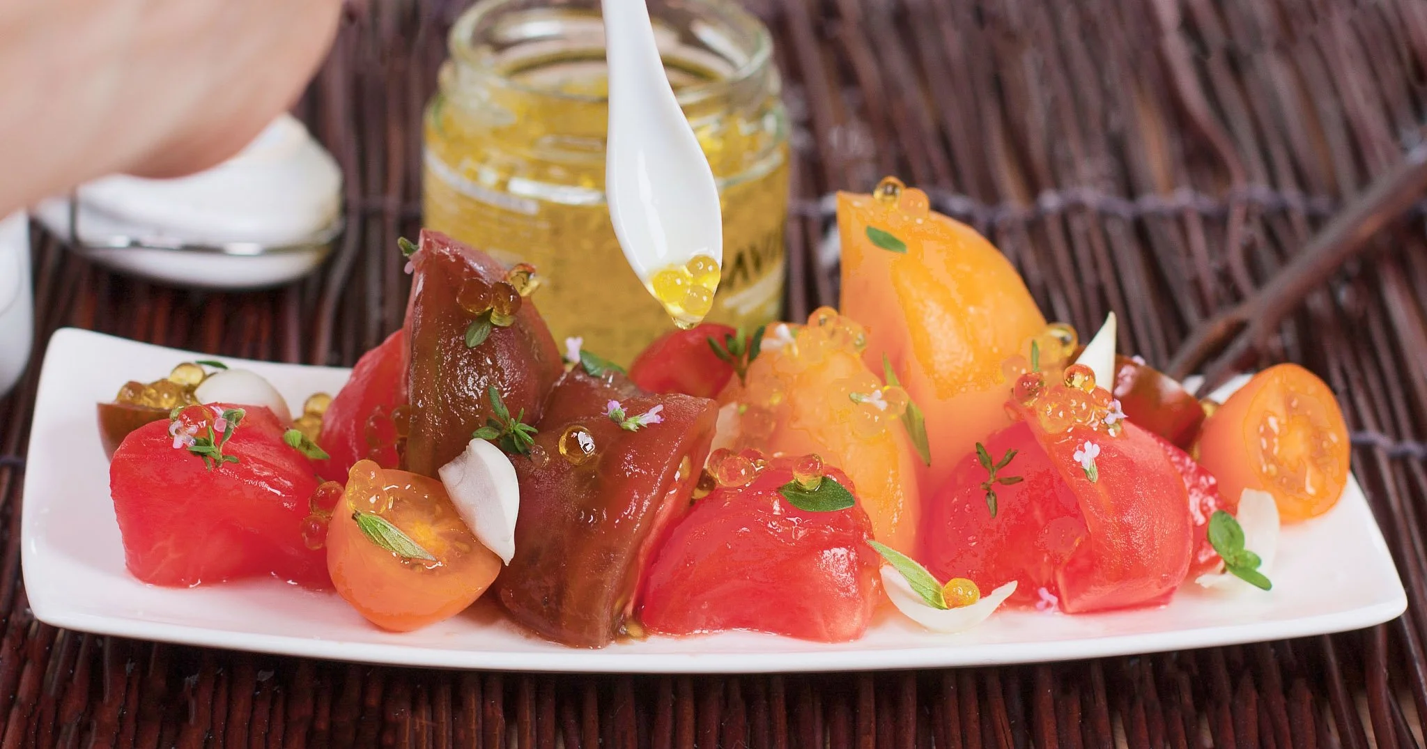 A white rectangular plate with watermelon and cantaloupe melons, topped with microgreens, edible flowers, and small dollops of white yogurt, garnished with a jar of honey in the background and a spoon with honey dripping onto the melons, set on a wov