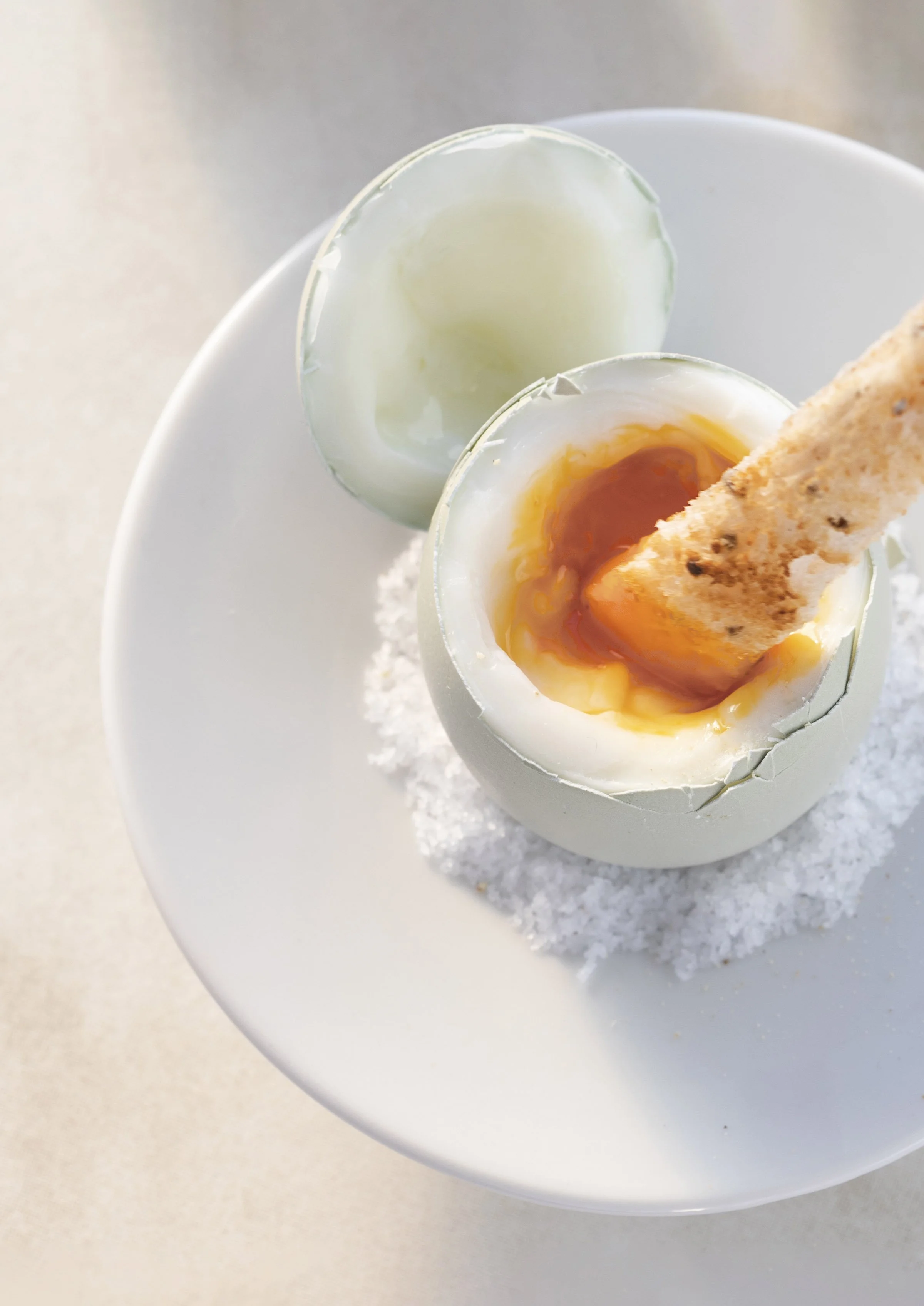 Soft-boiled egg in an eggshell on a white plate, with the top removed, revealing a runny yolk and a breadstick inside, garnished with salt or seasoning.