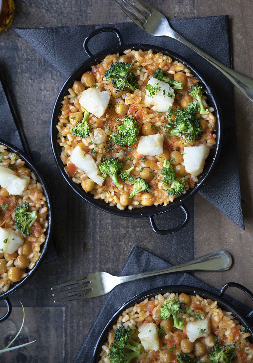 Baked chickpea and rice casserole topped with broccoli, cauliflower, and shredded cheese served in black oval dishes, with forks on black napkins.