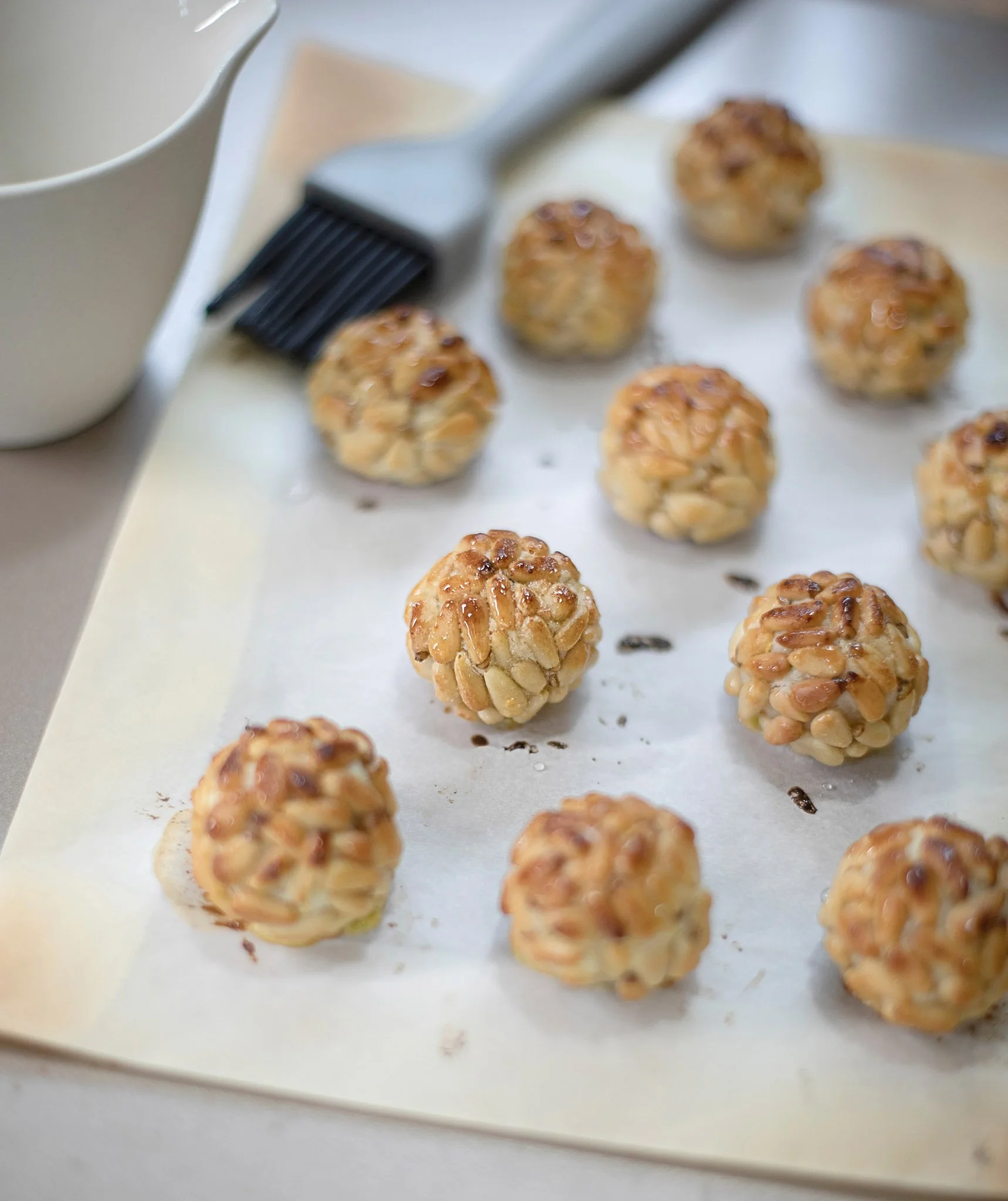 Tray of baked dessert balls covered in chopped nuts on parchment paper with a pastry brush and a small white bowl nearby.