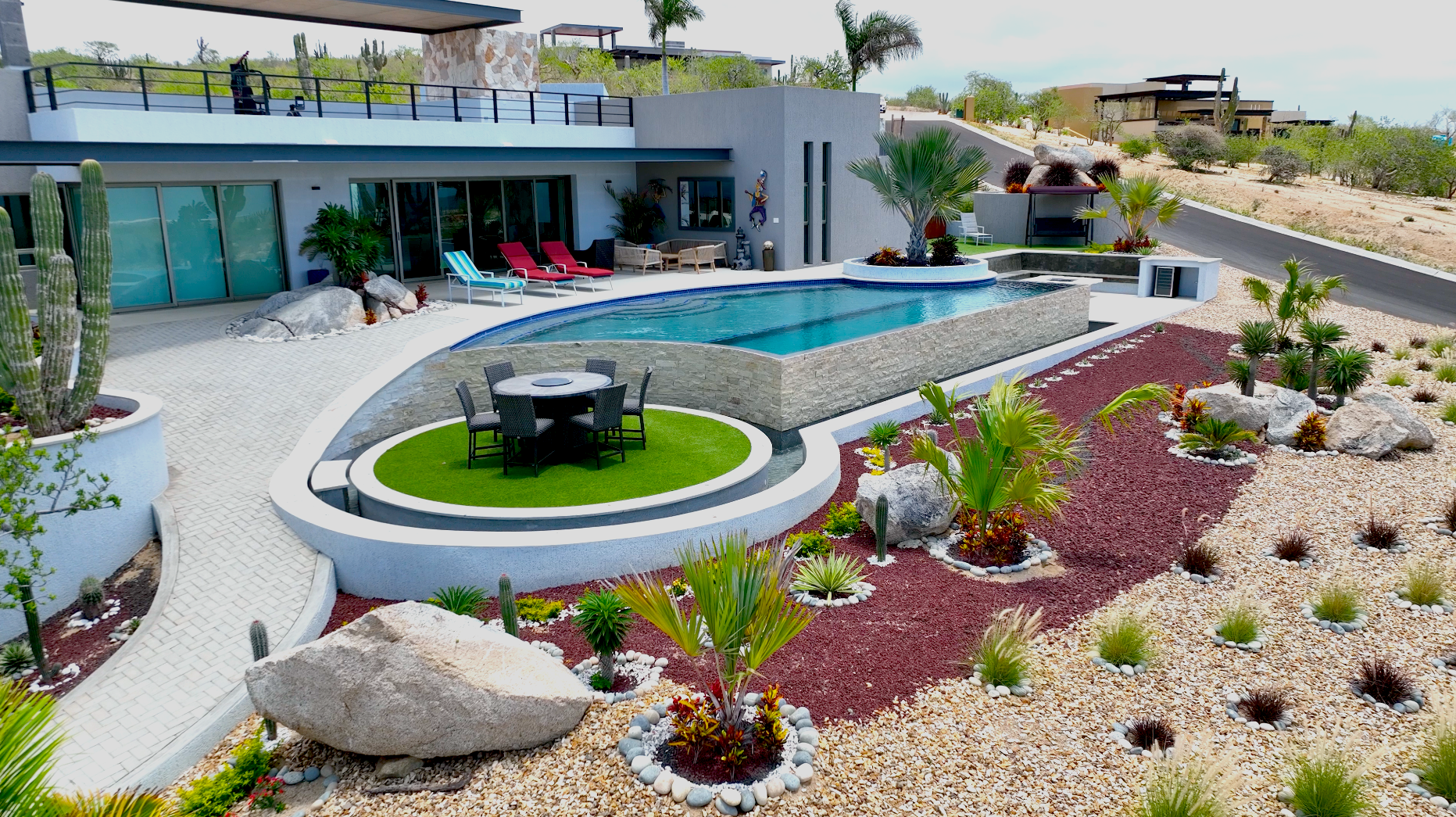 Drone aerial of a modern desert villa featuring a freeform pool, artificial turf, decorative rock garden, rooftop terrace, and native cactus landscaping