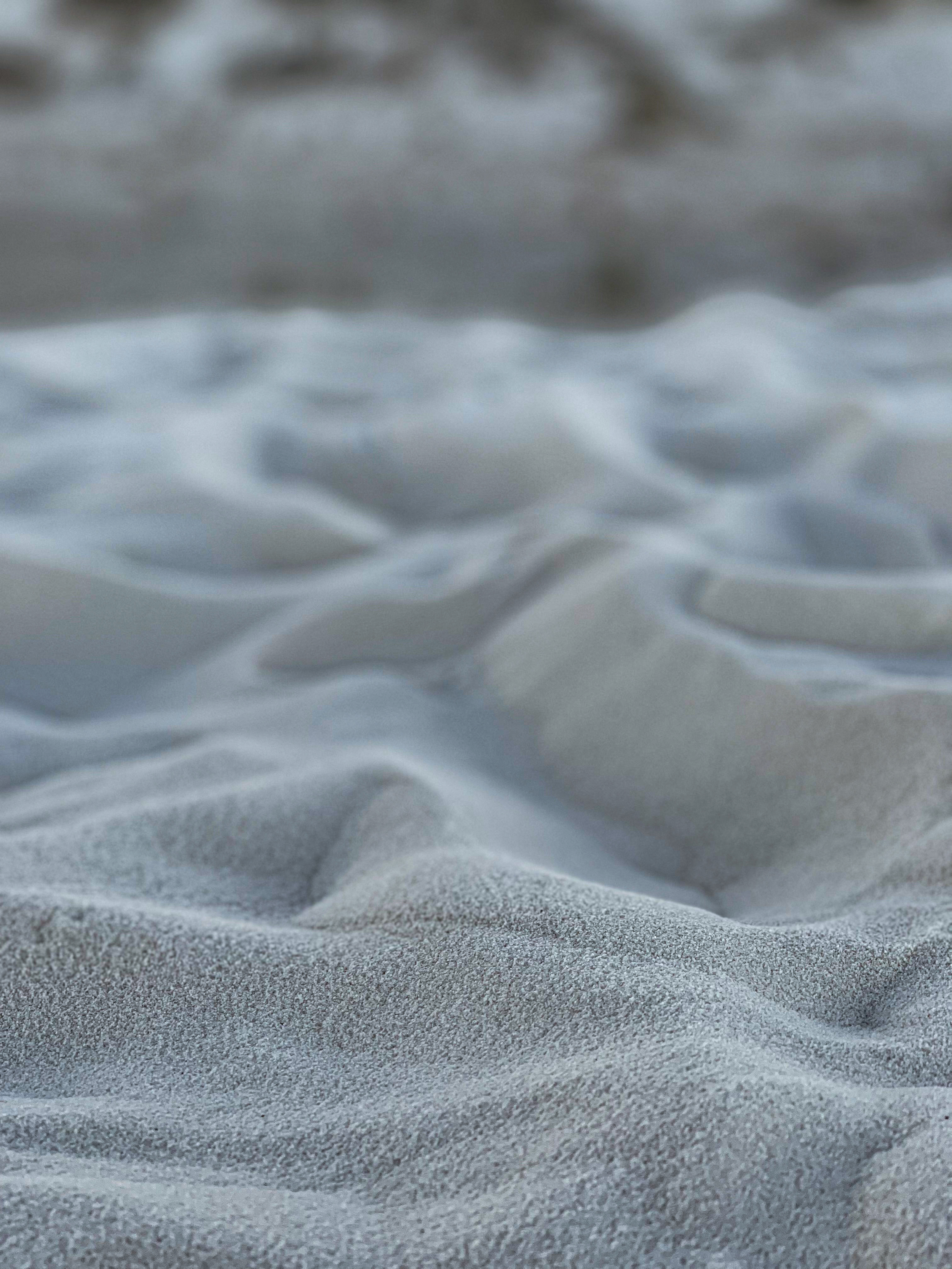 Close-up of sandy beach with textured sand dunes, blurred ocean waves in the background.