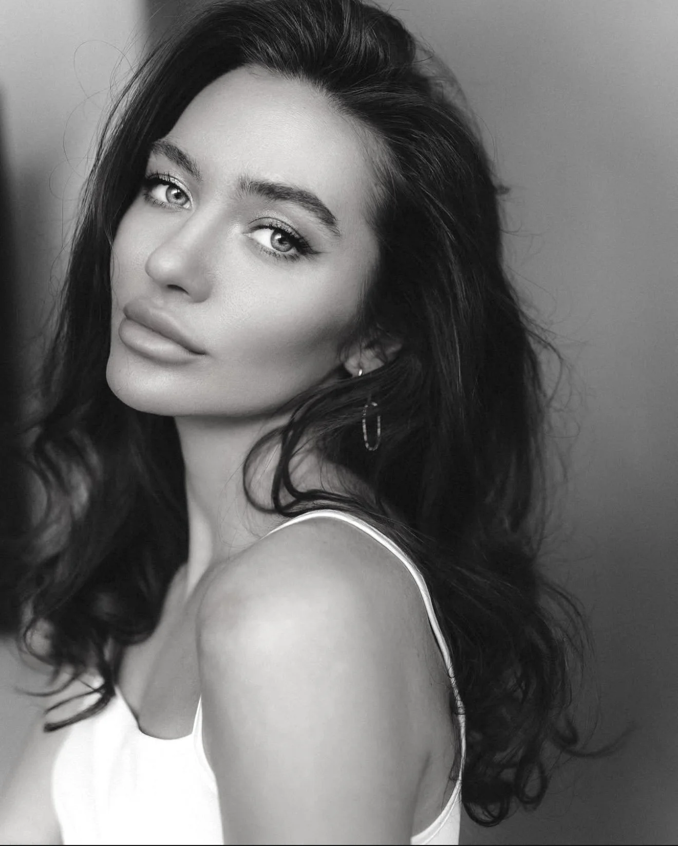 Black and white portrait of a woman with wavy dark hair, light-colored eyes, and full lips, wearing a sleeveless top and a small earring.