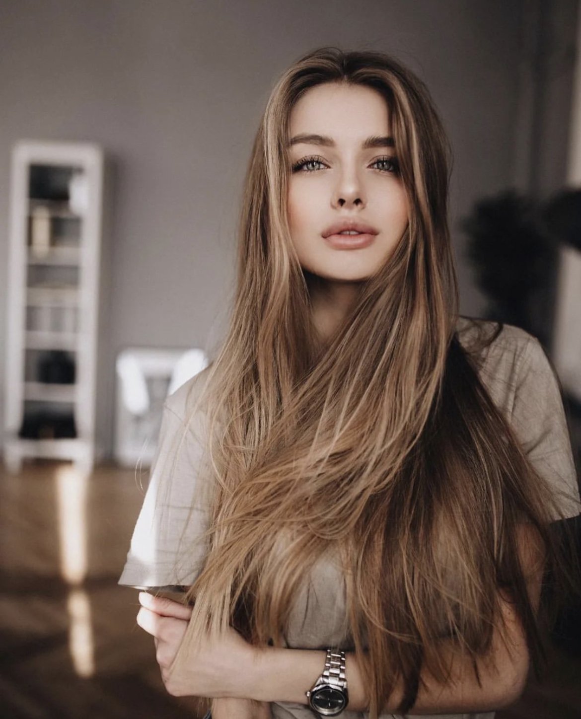 A young woman with long, wavy, light brown hair, wearing a beige top and a silver watch, standing in a room with blurred furniture in the background.