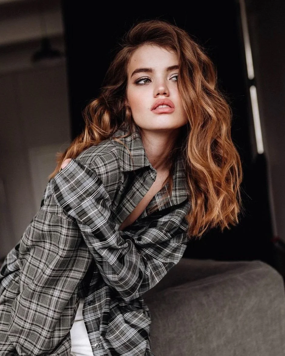 A young woman with long, wavy auburn hair, wearing a gray plaid shirt, looks to the side with a thoughtful expression, sitting in a dimly lit room.