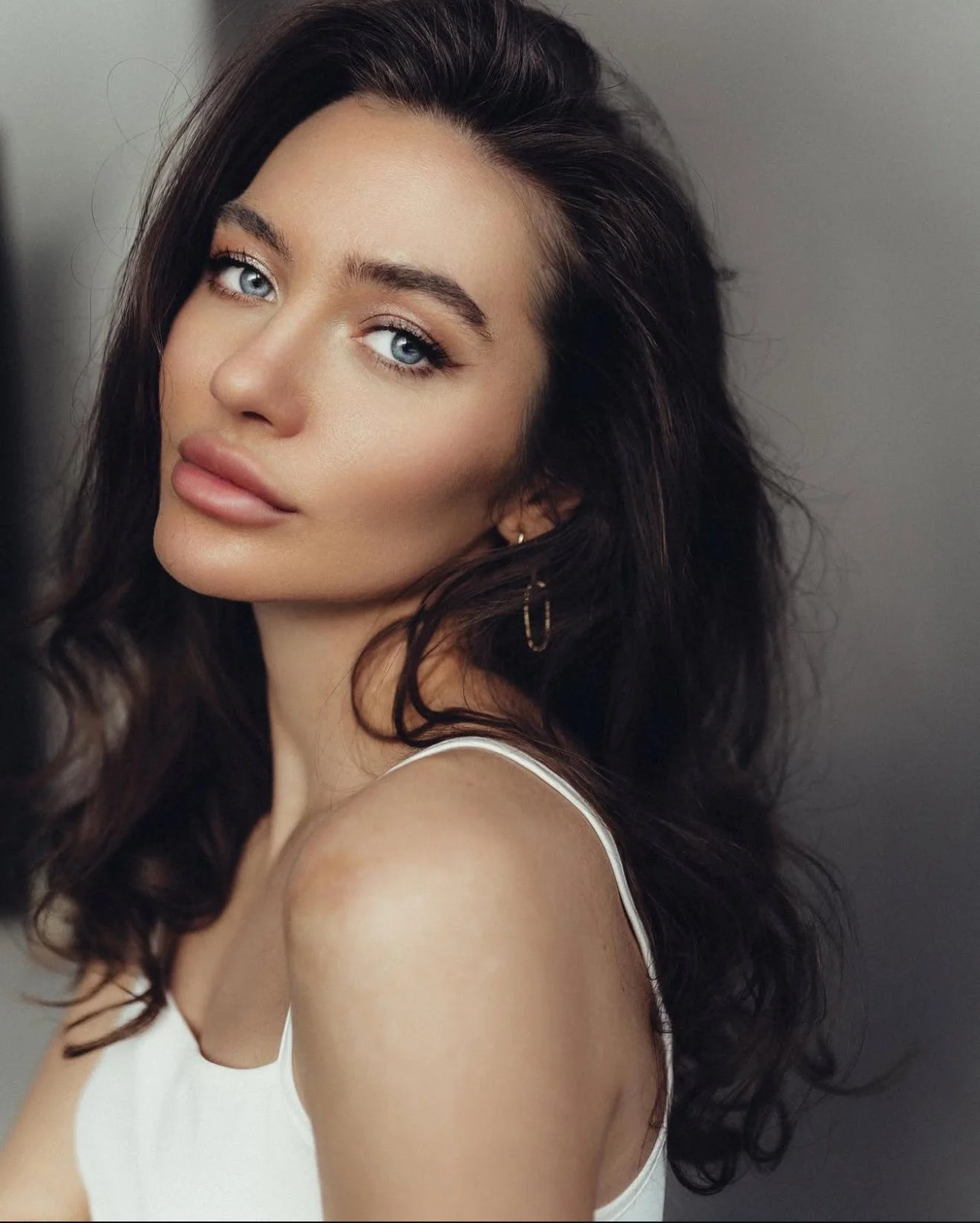 A young woman with long, wavy dark hair, blue eyes, and full lips, wearing a white top and gold hoop earrings, looking at the camera against a plain background.