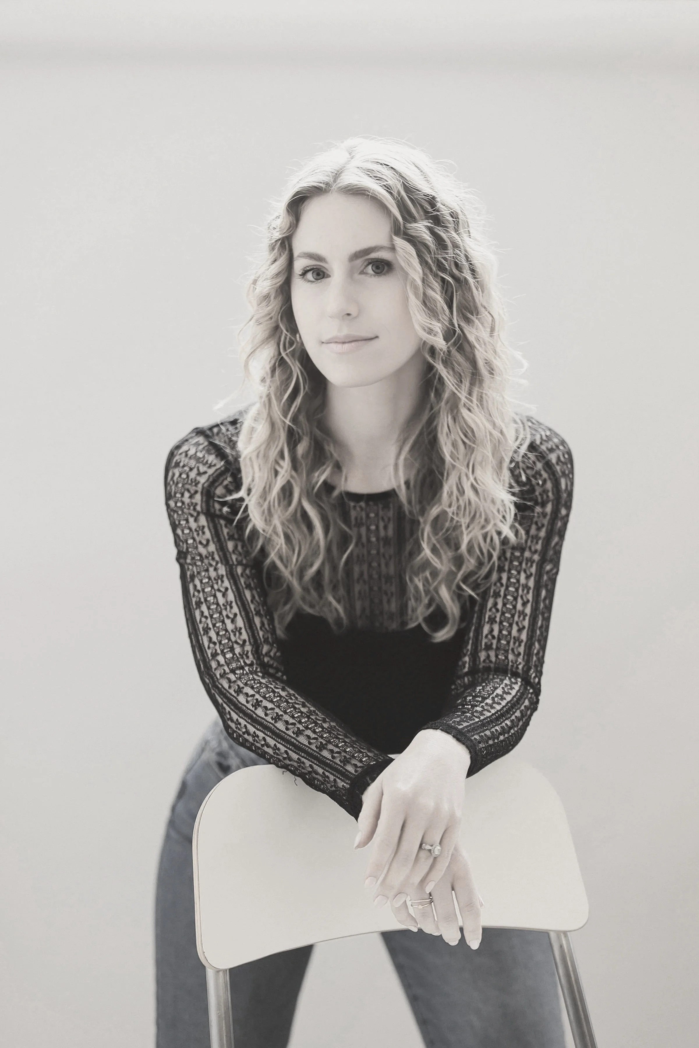 Black and white portrait of a young woman with curly hair, wearing a lace long-sleeve top, holding onto a white chair, against a plain background.