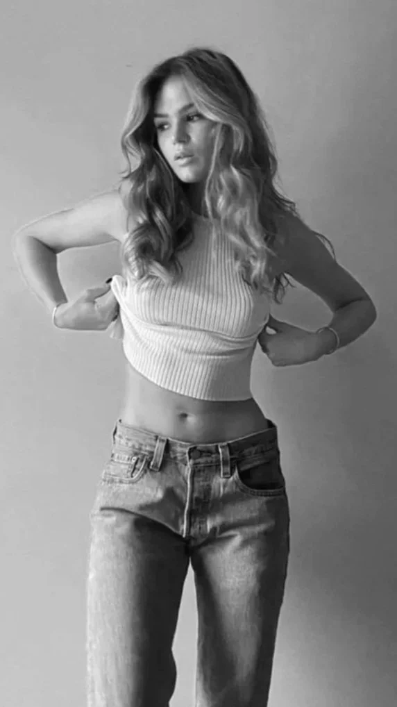 Black and white photo of a young woman with wavy hair, wearing a sleeveless crop top and high-waisted jeans, standing against a plain background with her hands on her hips.