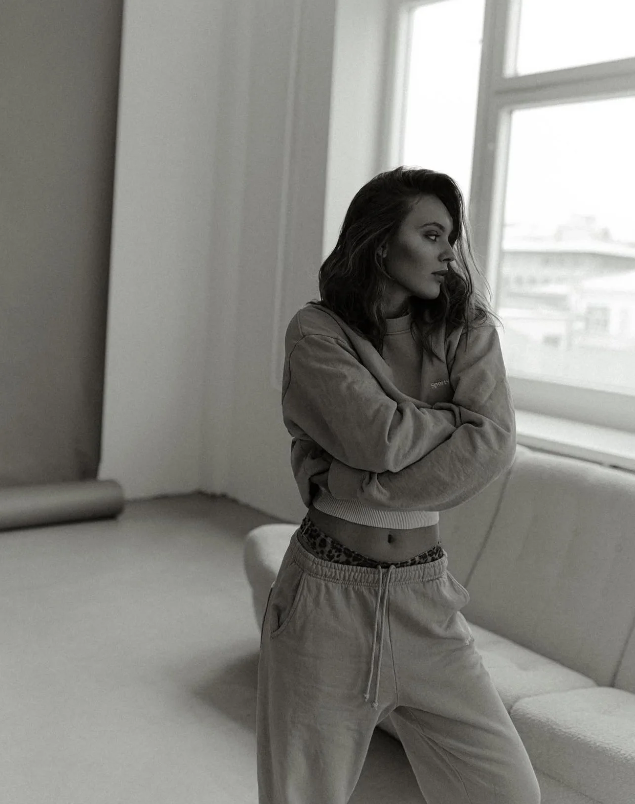 A young woman with dark, wavy hair standing indoors near a window with a city view, wearing a cropped sweatshirt and sweatpants, with her arms crossed.