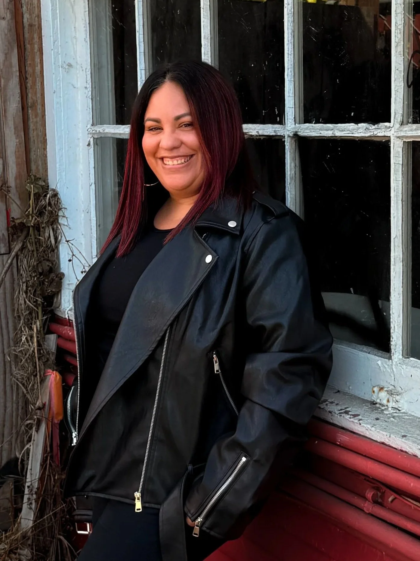 A young woman with shoulder-length dark hair with red highlights, smiling and wearing a black leather jacket, standing by a weathered window with peeling paint and a red exterior wall.