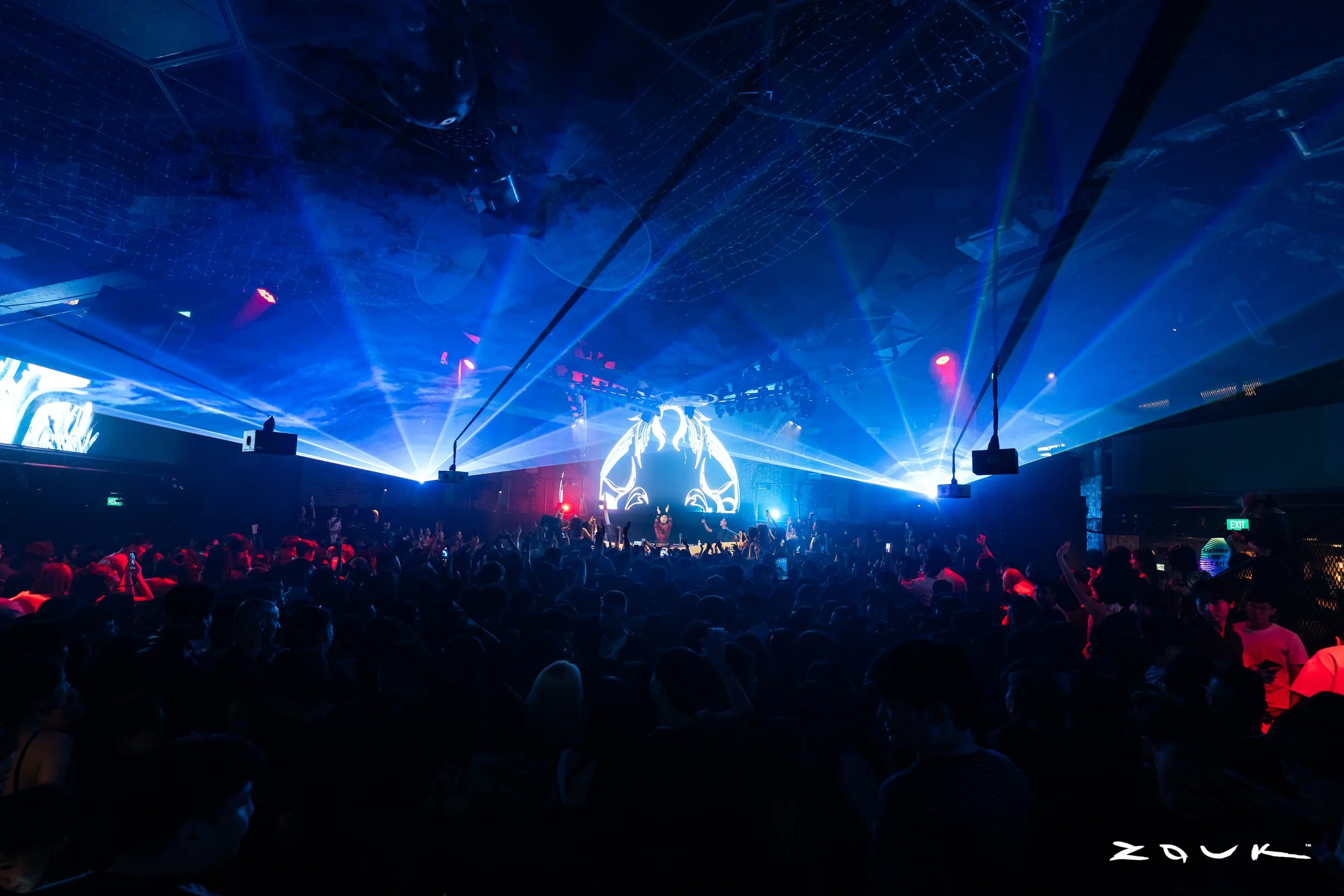 DJ Wukong performing live at Zouk Singapore as blue laser beams illuminate a packed crowd, creating an immersive nightclub atmosphere during a high-energy electronic music performance.