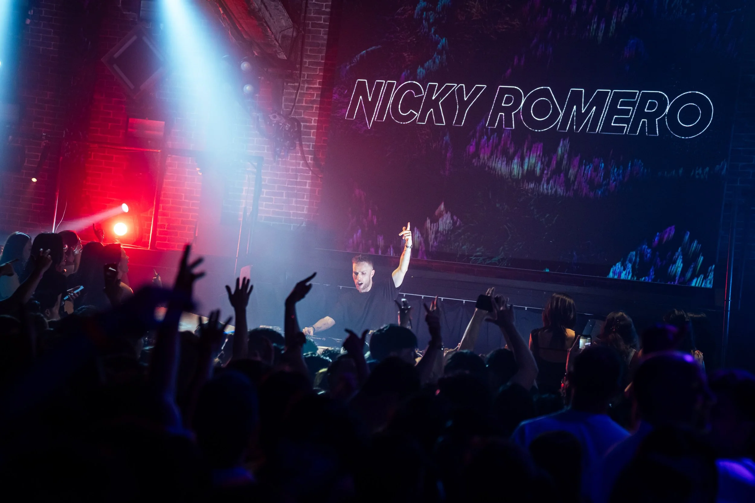 DJ Nicky Romero performing live at Zouk Singapore, engaging a packed crowd beneath dramatic blue and red stage lighting with immersive LED visuals during a high-energy electronic dance music performance.