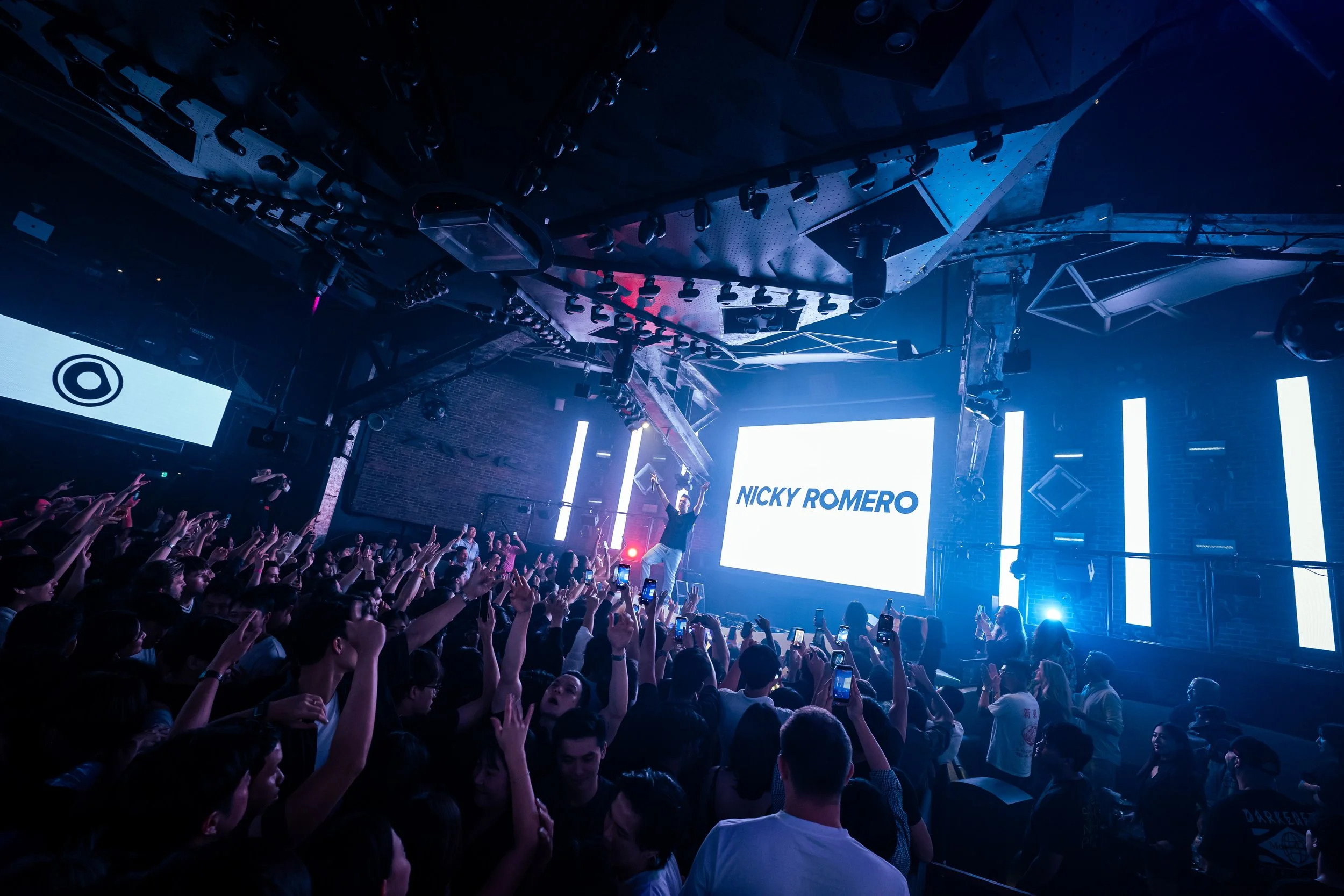 DJ Nicky Romero performing live at Zouk Singapore as a packed crowd raises their hands beneath immersive LED visuals and vibrant blue stage lighting during a high-energy electronic dance music performance.
