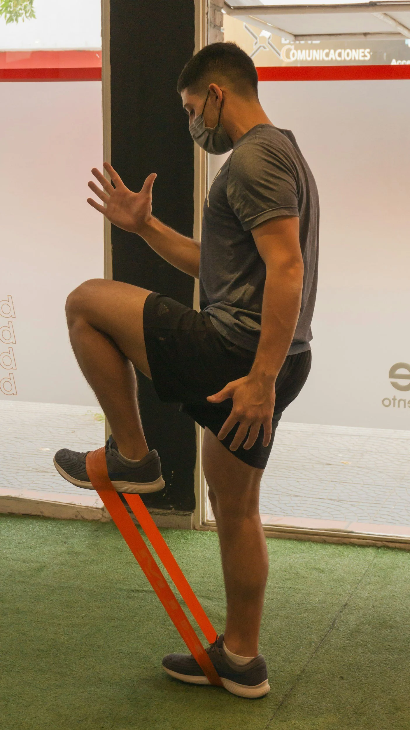 A man wearing a face mask and workout clothes is balancing on one leg while using an orange resistance band around his ankle for exercise. He is inside near a doorway.