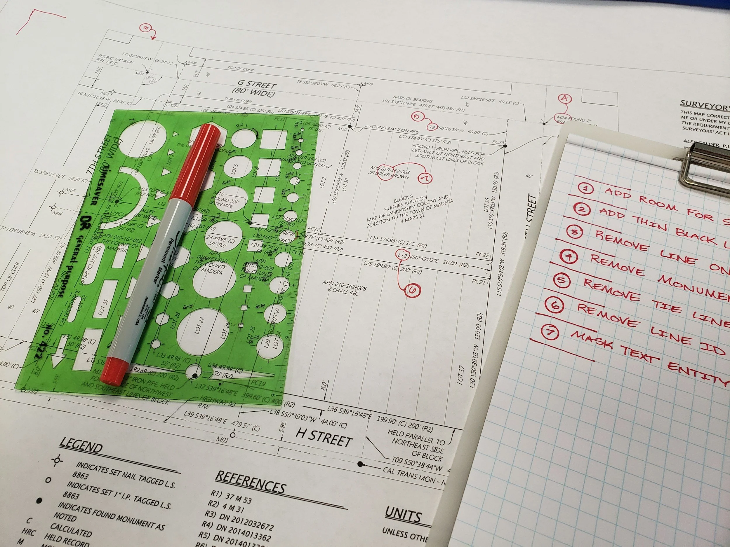 A blueprint with a green plastic stencil and red marker on top, and a notepad with numbered instructions written in red ink.