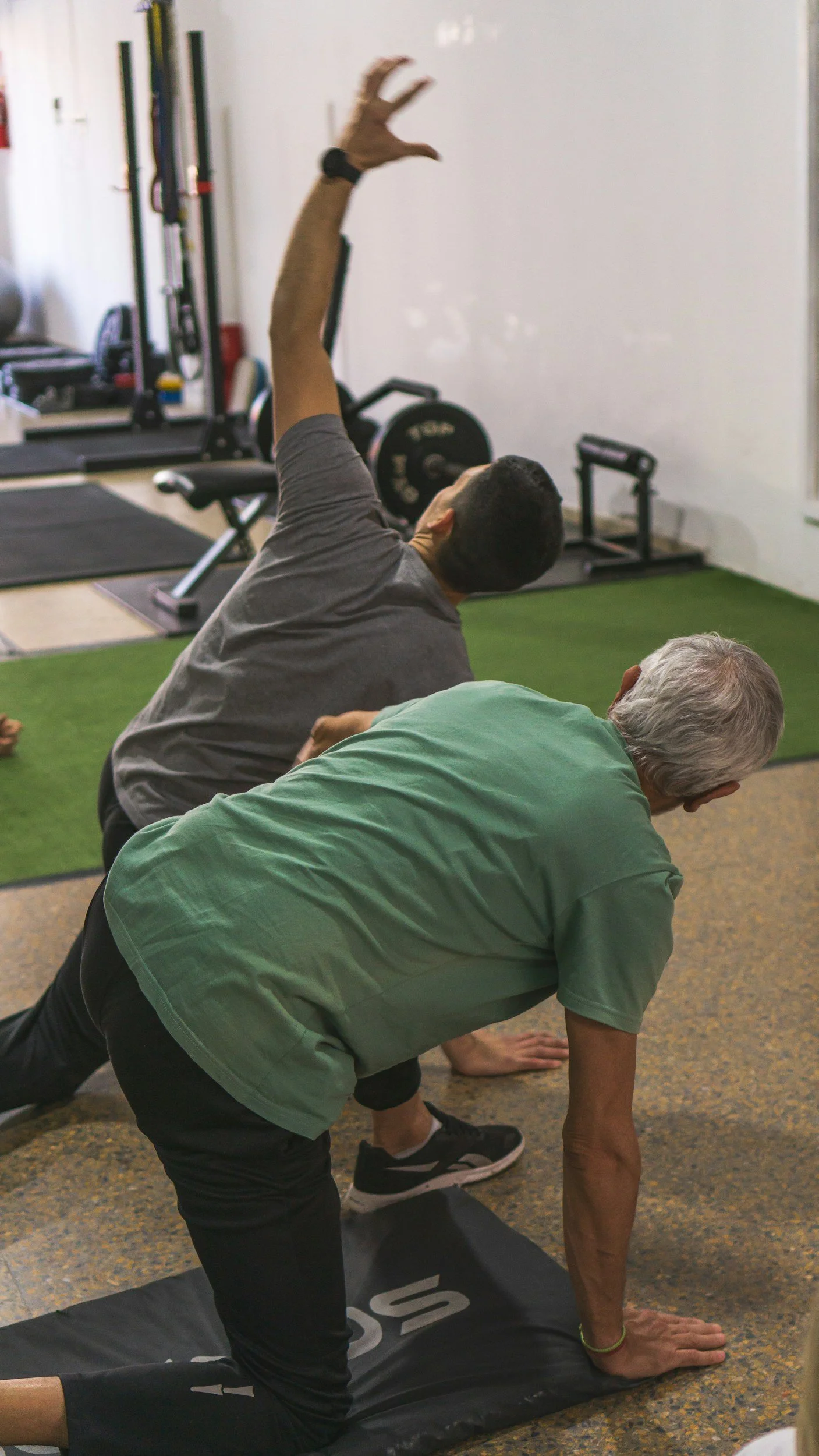 A man with gray hair practicing yoga on a black mat while a younger man in a gray shirt performs a yoga pose nearby in a gym with weightlifting equipment in the background.
