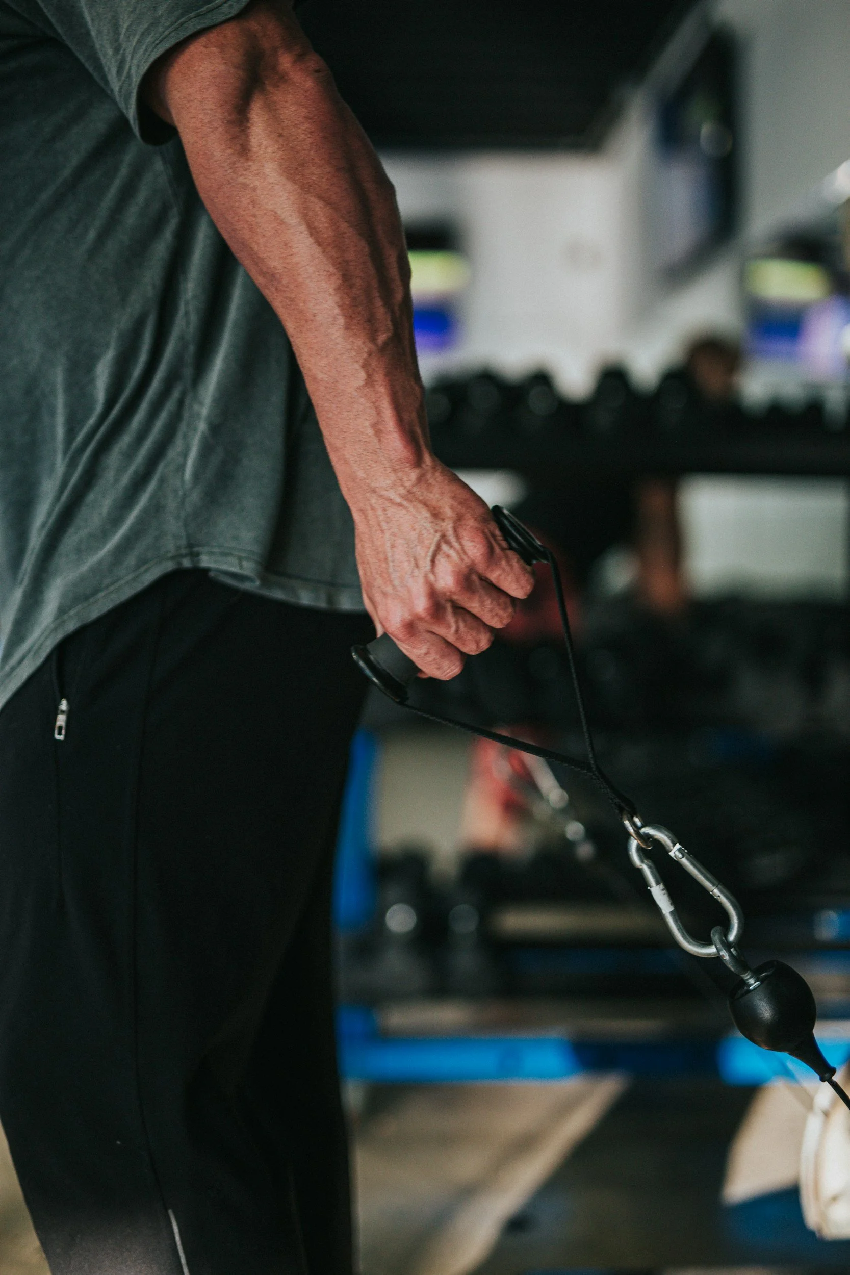 Close-up of a person holding a black speed bag with a lanyard attached inside a gym. The person's arm and hand are visible, wearing a grey shirt and black pants.