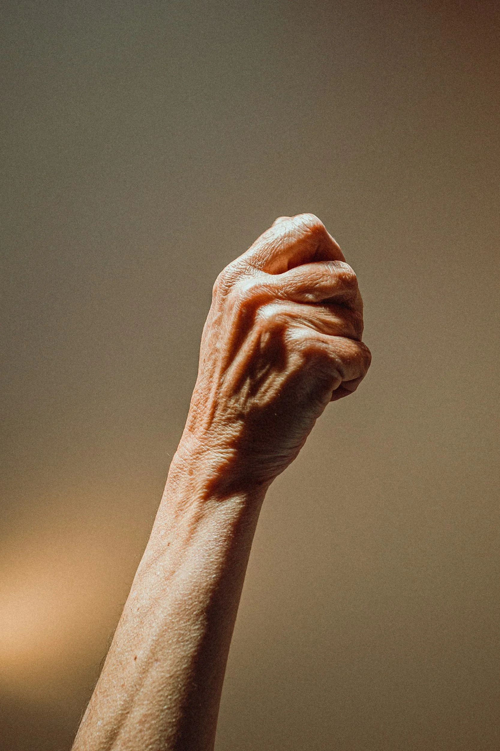 Raised fist of an elderly person with visible veins and wrinkles against a plain background.