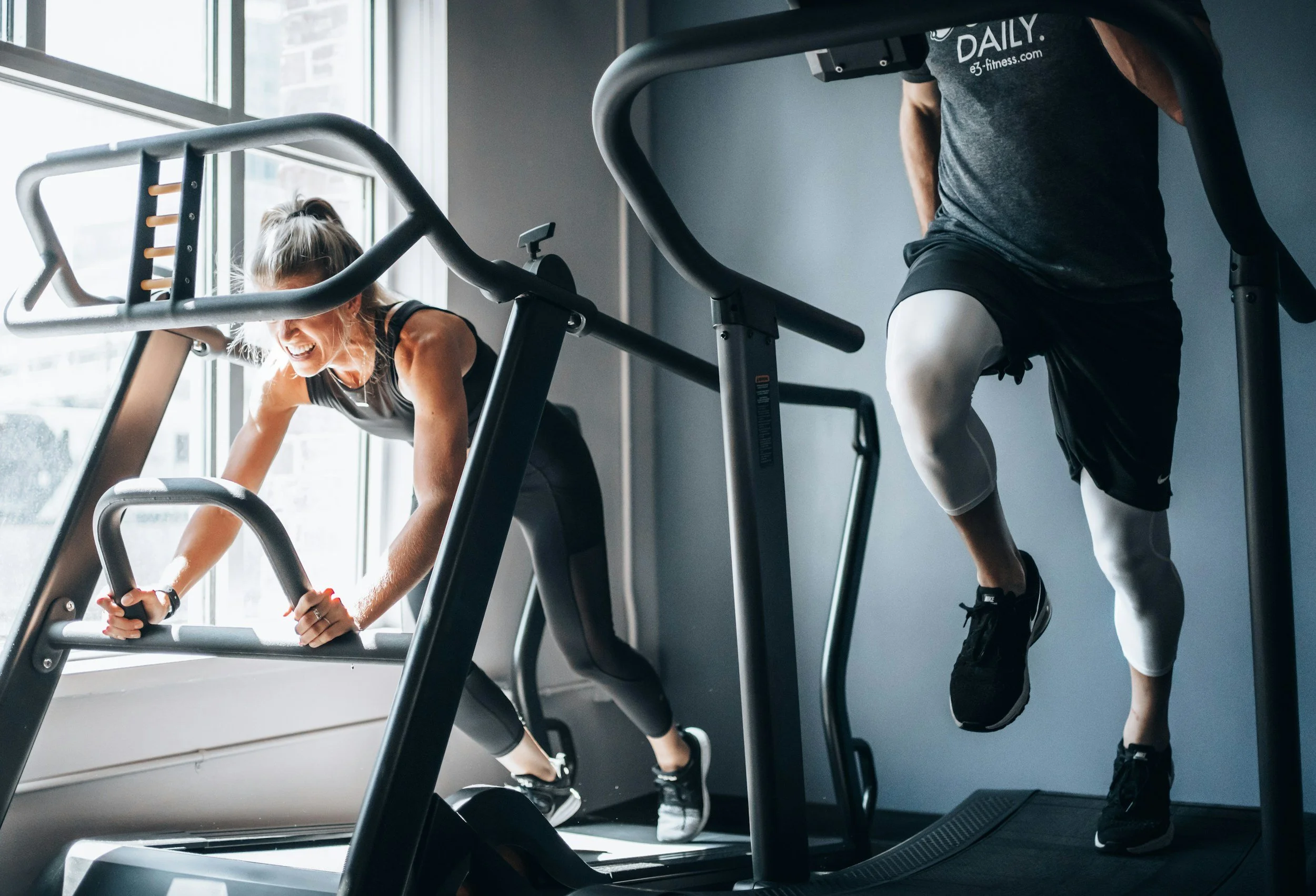 A woman and a man working out on treadmills in a gym, with a woman leaning forward holding the treadmill handlebars and smiling, and a man doing a step-up exercise on the side.