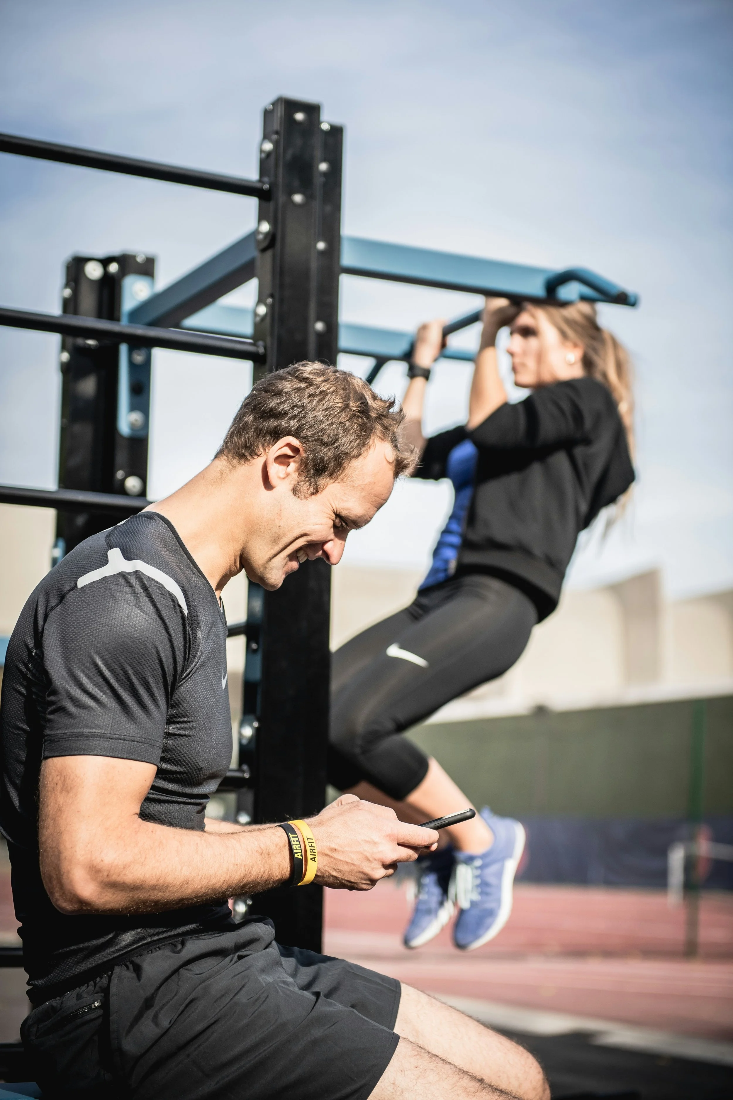 A man sitting on the ground at an outdoor fitness area, looking at his phone, with a woman doing pull-ups on a bar behind him.