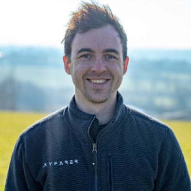 Smiling man outdoors wearing a black jacket with a blurred landscape background.
