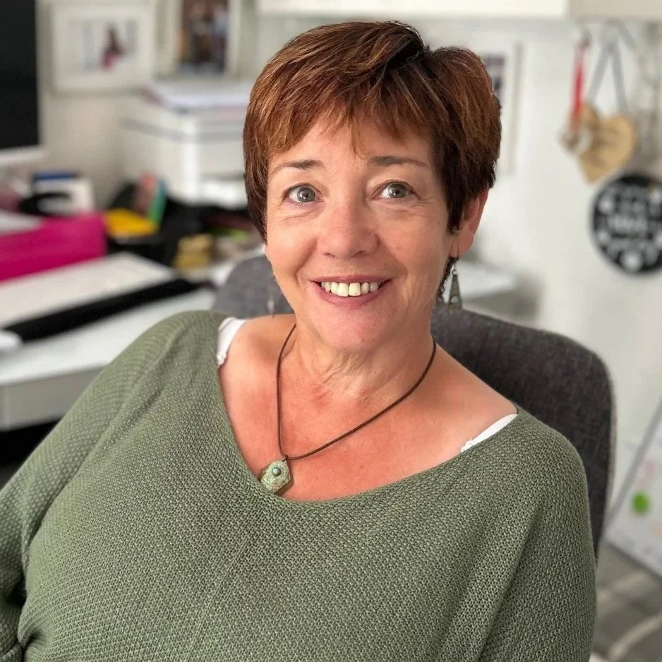 A woman with short brown hair and a bright smile, wearing a green top and a necklace, sitting in an office environment.