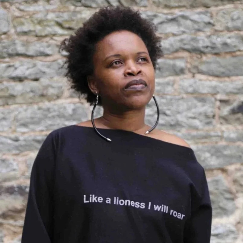 A woman with natural curly hair wearing large hoop earrings and a black shirt with white text that reads "Like a lioness I will roar," standing in front of a stone wall.