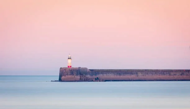 Coastal lighthouse on a rocky breakwater beside calm sea, representing the Passage open call by Sussex Contemporary.