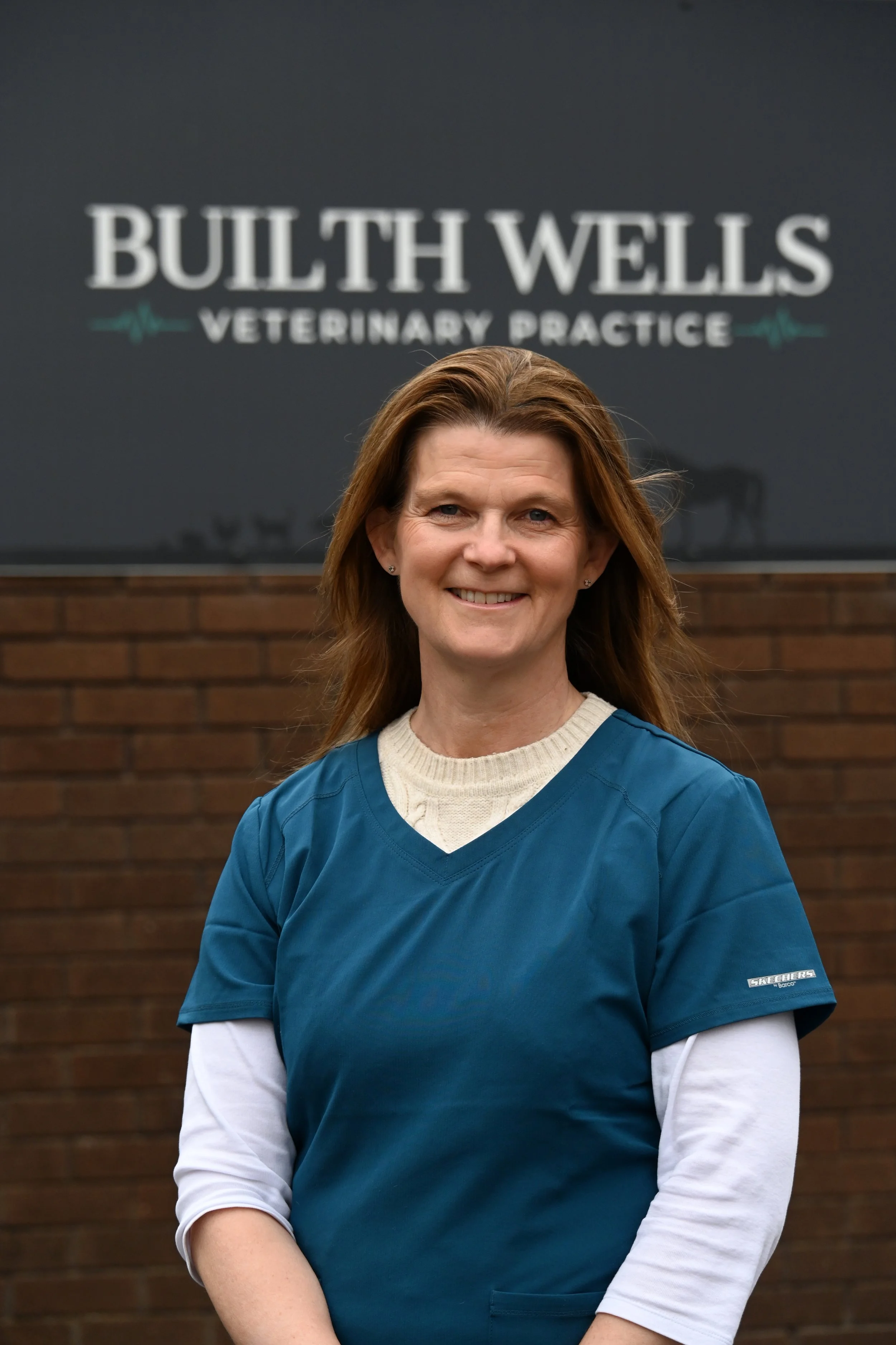 A woman standing in front of a sign that reads "BULTH WELLS VETERINARY PRACTICE." The woman is wearing a blue medical scrubs and smiling.