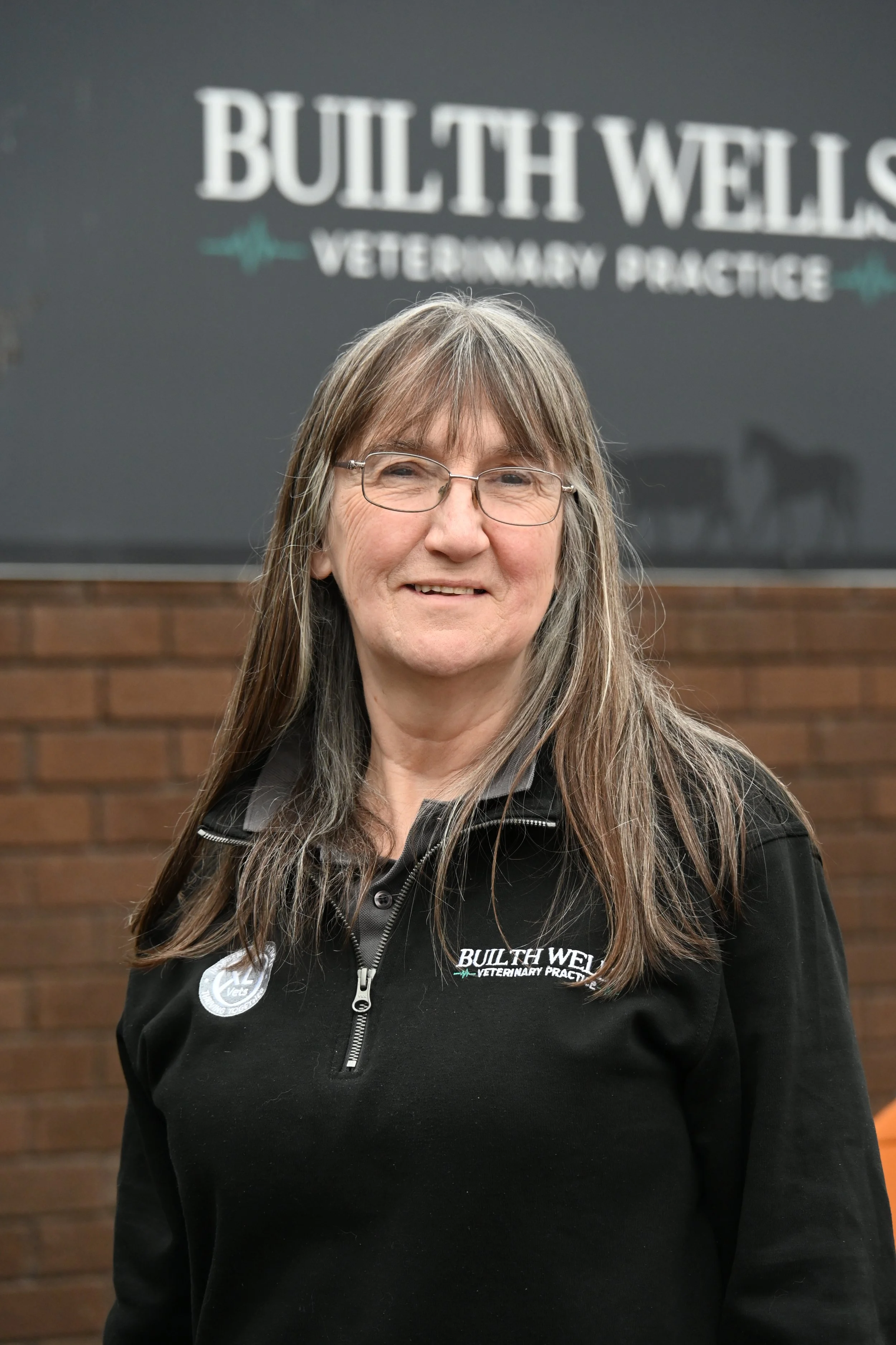 A woman with long brown hair, glasses, and a black jacket standing in front of a veterinary practice sign.