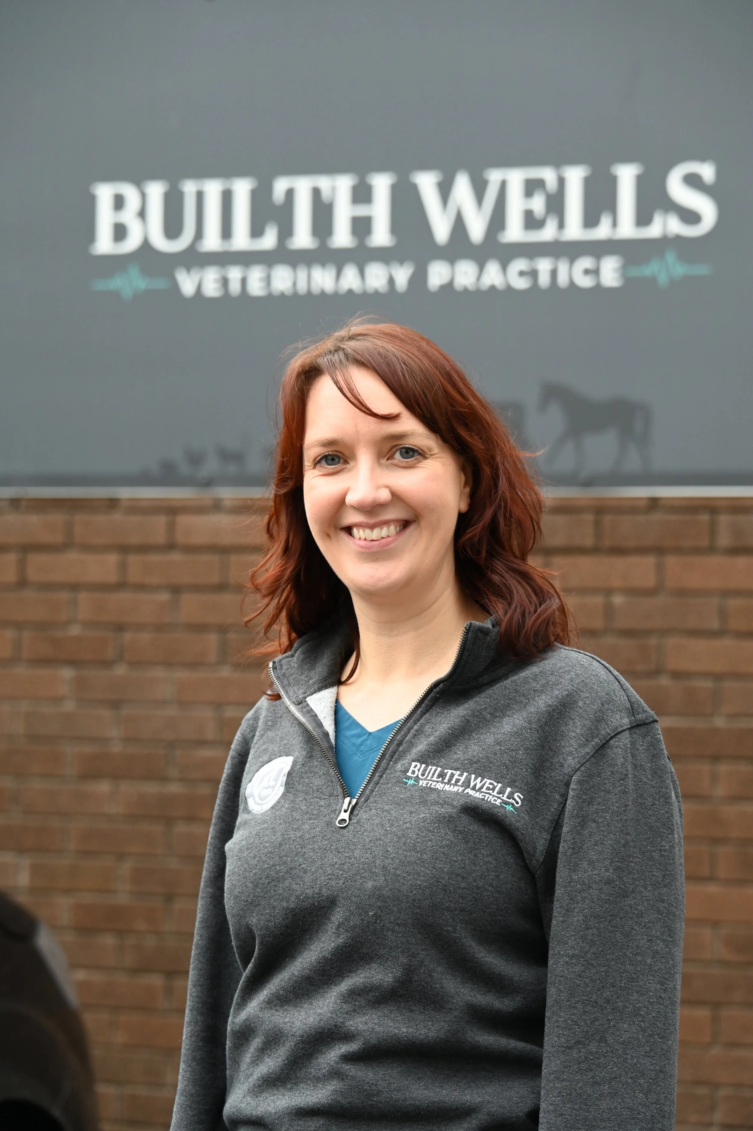 A woman smiling in front of a sign and brick wall, wearing a gray jacket with 'BULTH WELLS VETERINARY PRACTICE' logo.