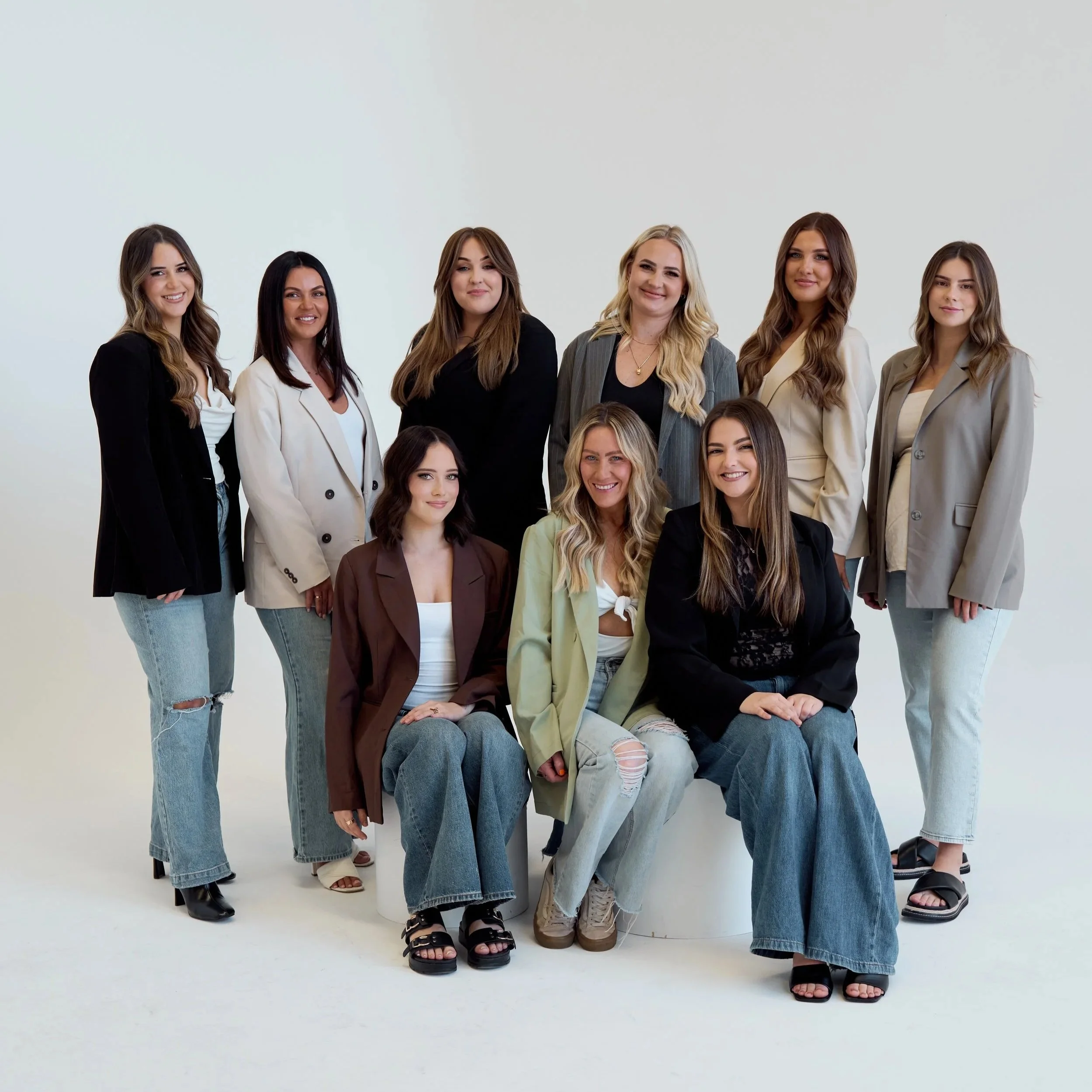 Group of nine hairstylists posing together against a plain white background, dressed in casual and business casual attire, smiling at the camera.