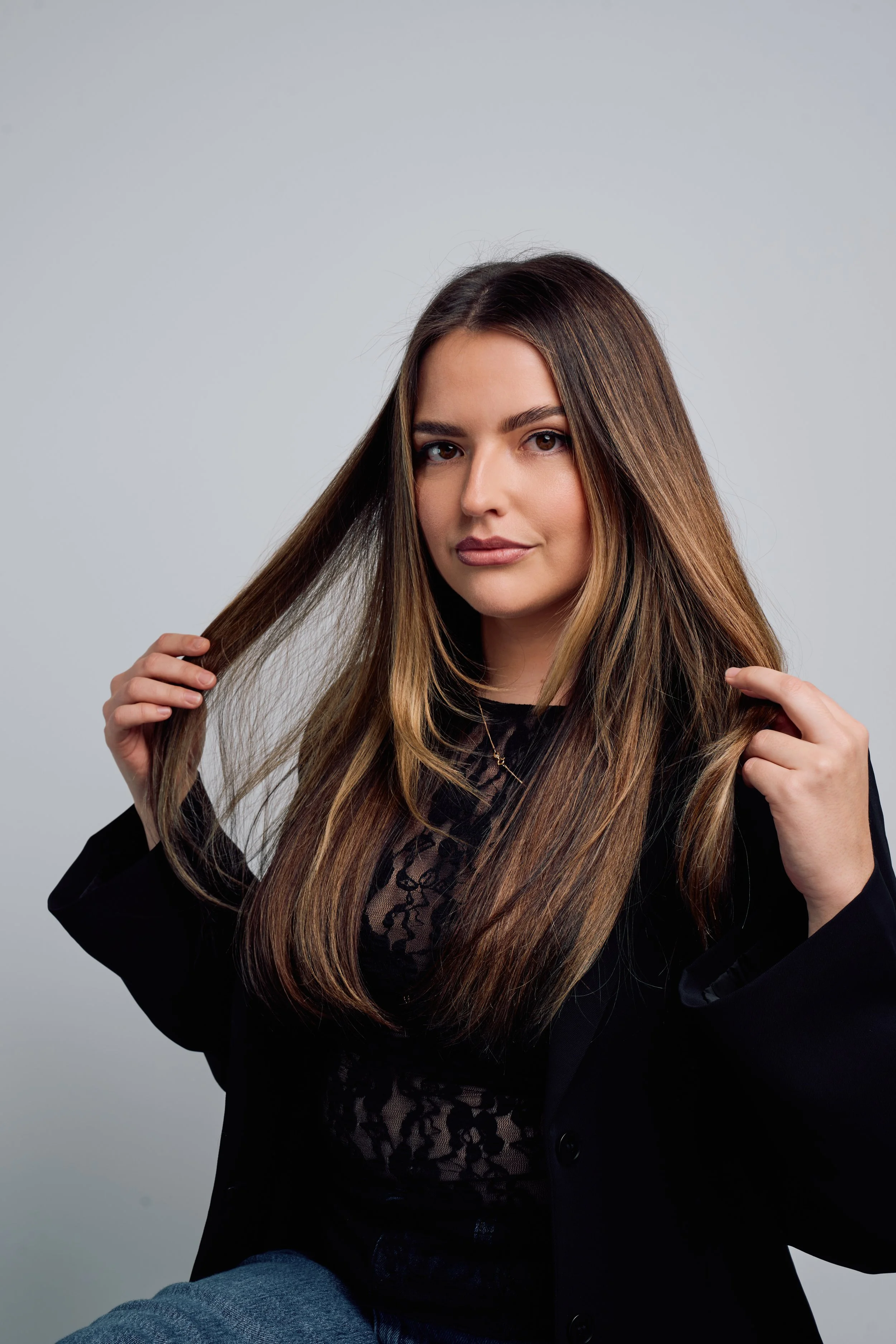 A marketing manager with long brown hair, holding strands of her hair with both hands, looking at the camera against a plain gray background.