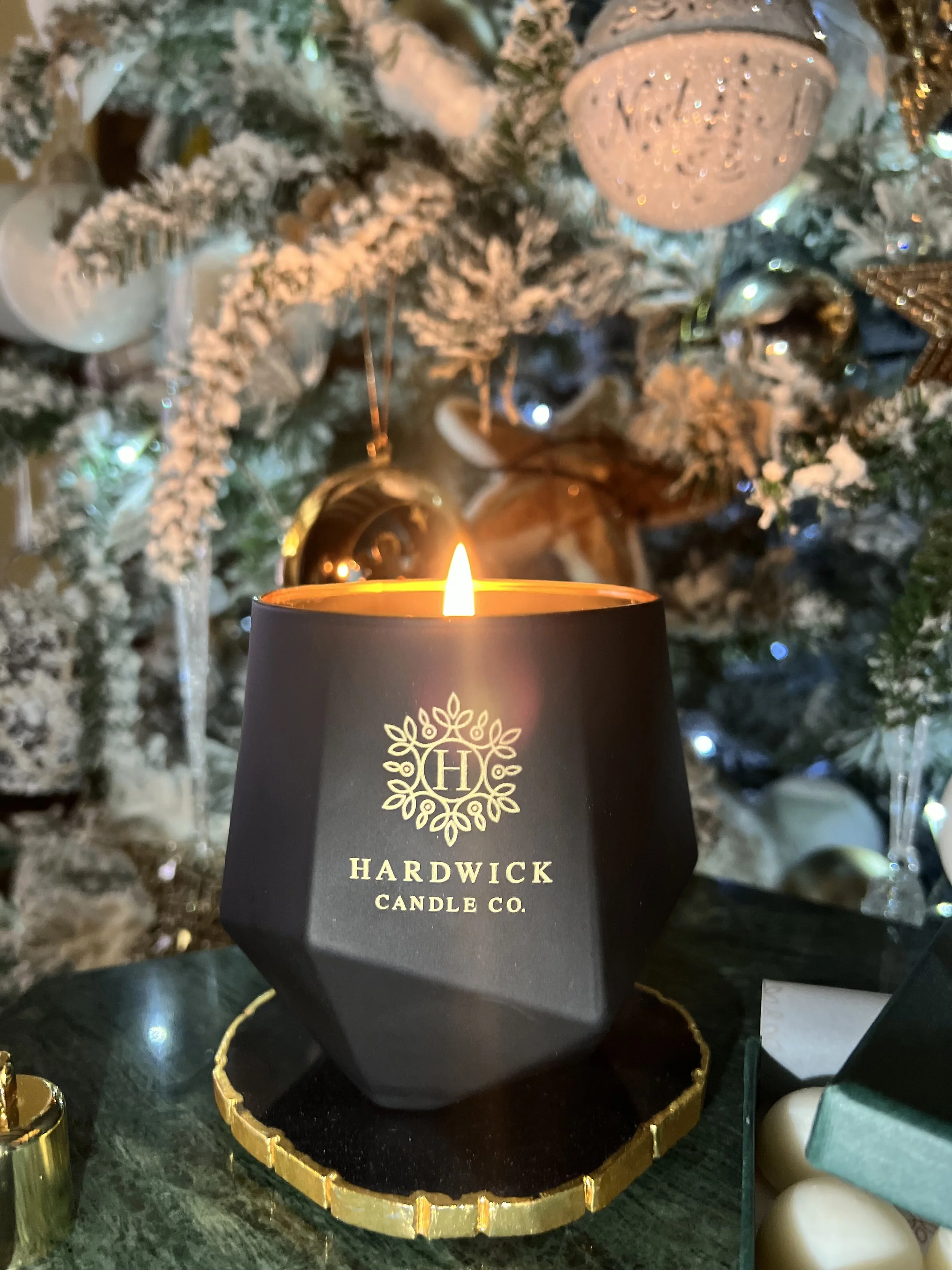 A lit black candle with gold lettering and a decorative logo reading 'Hardwick Candle Co.' is placed on a black coaster with gold accents. In the background, part of a decorated Christmas tree with snow-like white ornaments, gold balls, and ornaments is visible.