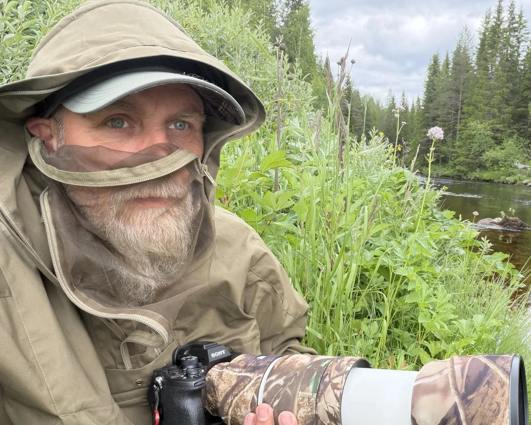En mand i udendørs tøj med kasket, jakke og camouflage-udstyr, der holder et kamera med teleobjektiv, omgivet af grøn vegetation nær en flod og fyrretræer.