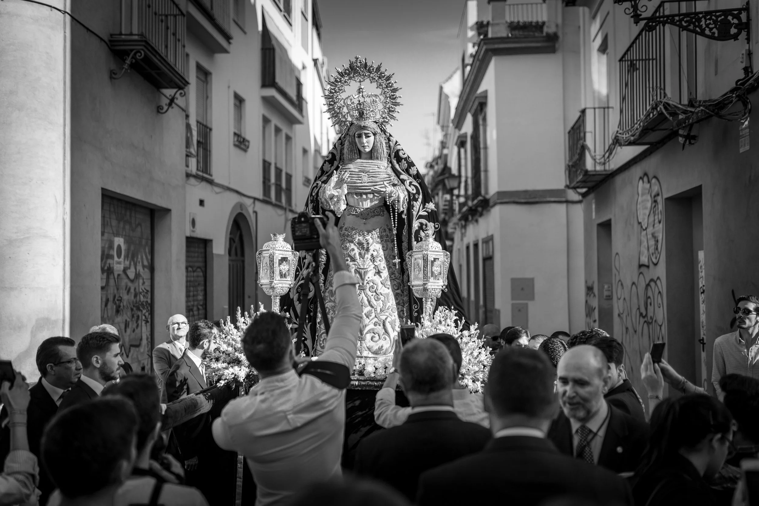 A religious procession on a city street with a large statue of a woman, likely a religious figure, adorned with a halo and robes. People gather and some take photos, with the scene in black and white.