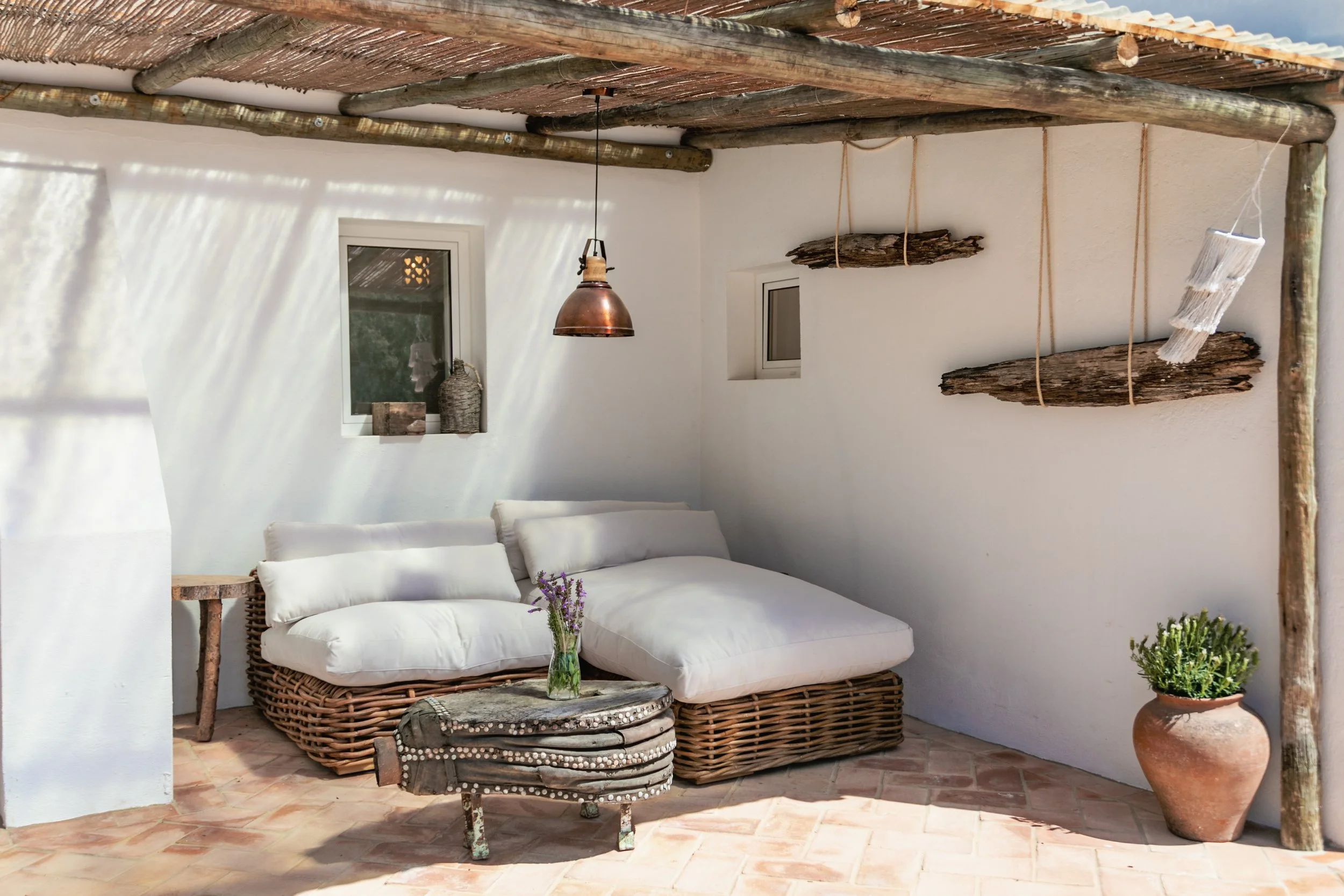 Outdoor patio with white walls, a wicker sofa with white cushions, a small rustic wooden table with a glass jar of lavender, a hanging copper light, wooden wall decor, pink tile flooring, and a large terracotta pot with greenery.