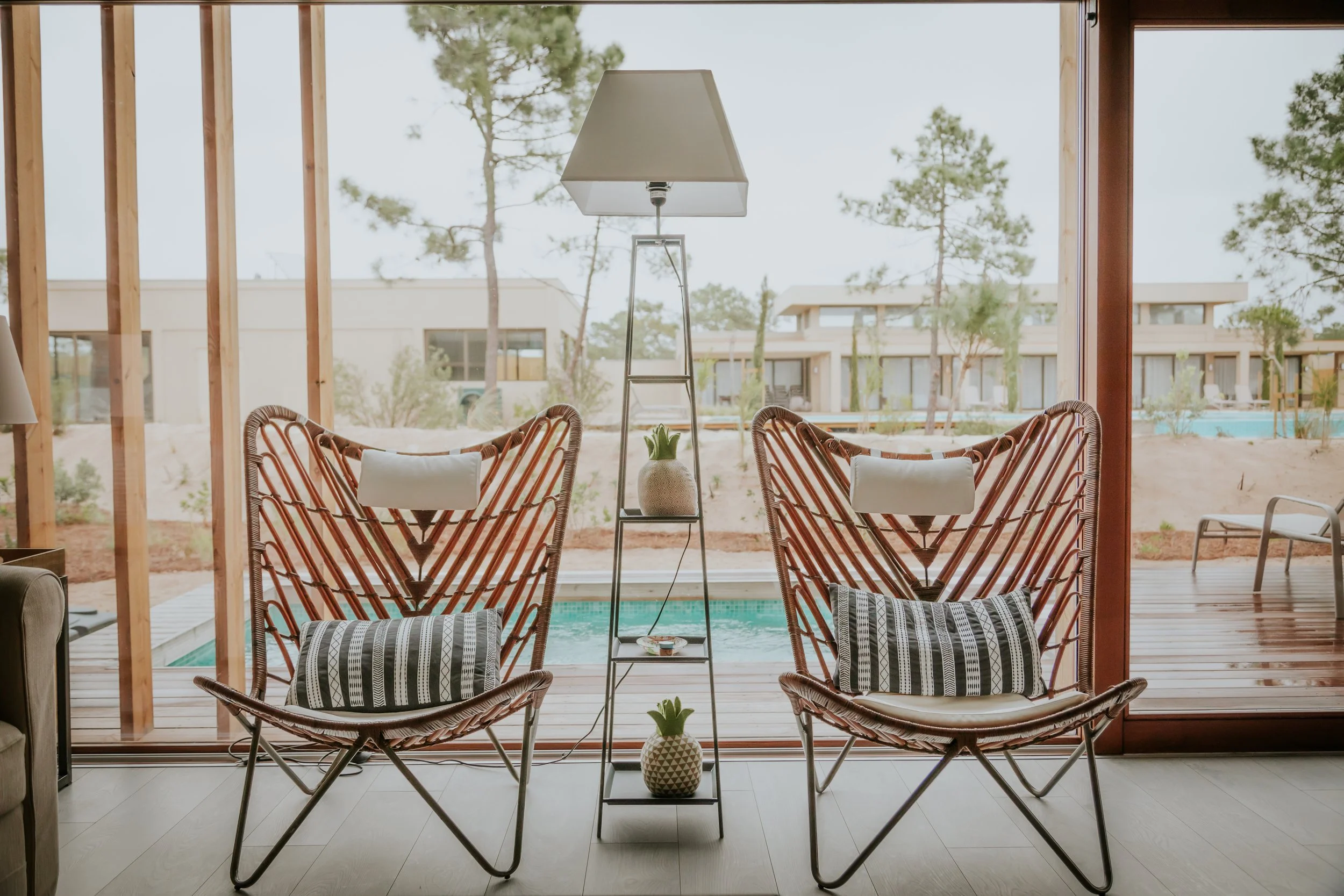 Interior view of two rattan chairs with cushions, a tall floor lamp, and a small shelf with potted plants, facing large glass doors opening to an outdoor patio with a pool and modern houses in the background.