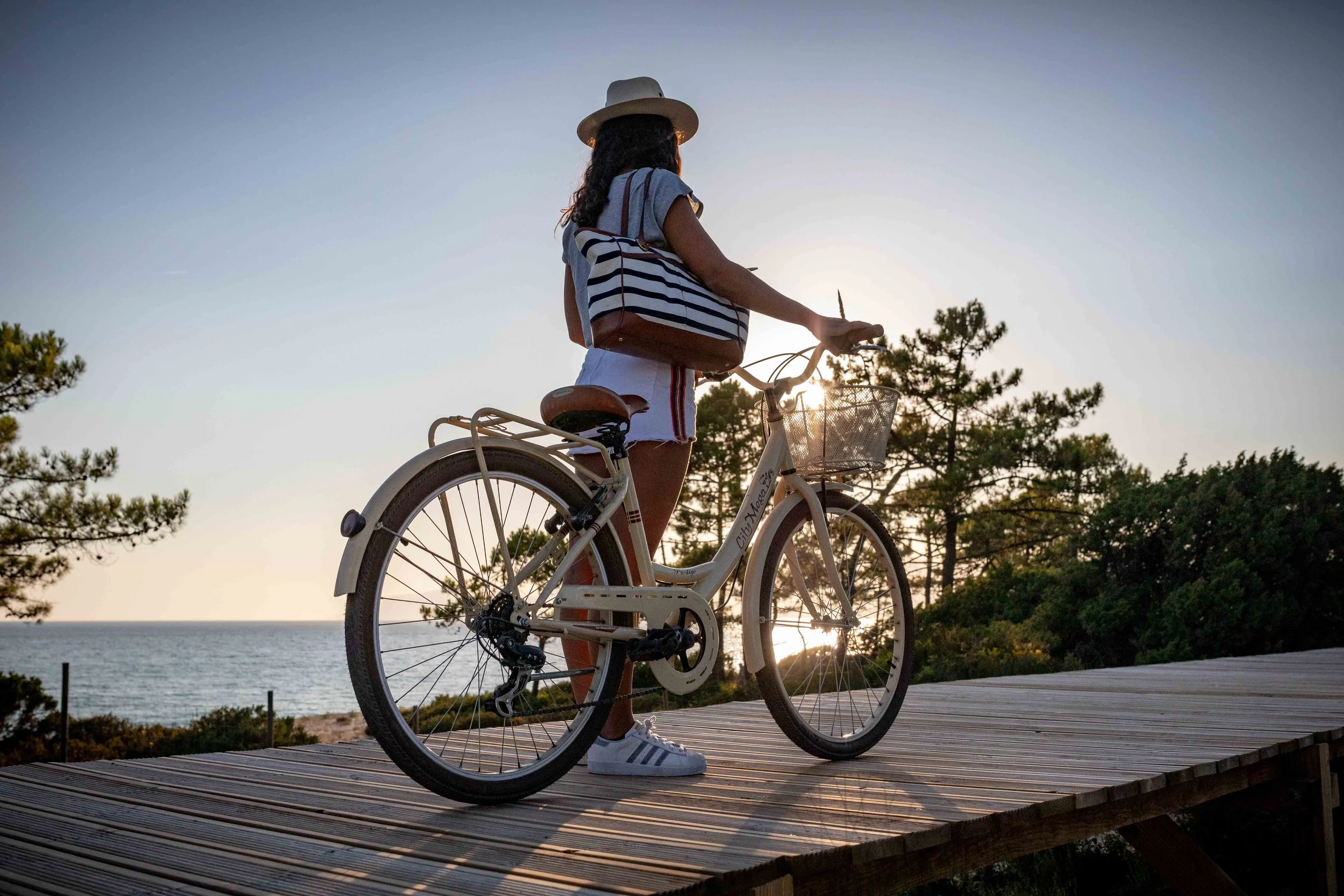 A woman with curly hair, wearing a hat, striped shirt, and shorts, stands with a bicycle on a wooden pathway by the beach during sunset, carrying a striped bag.
