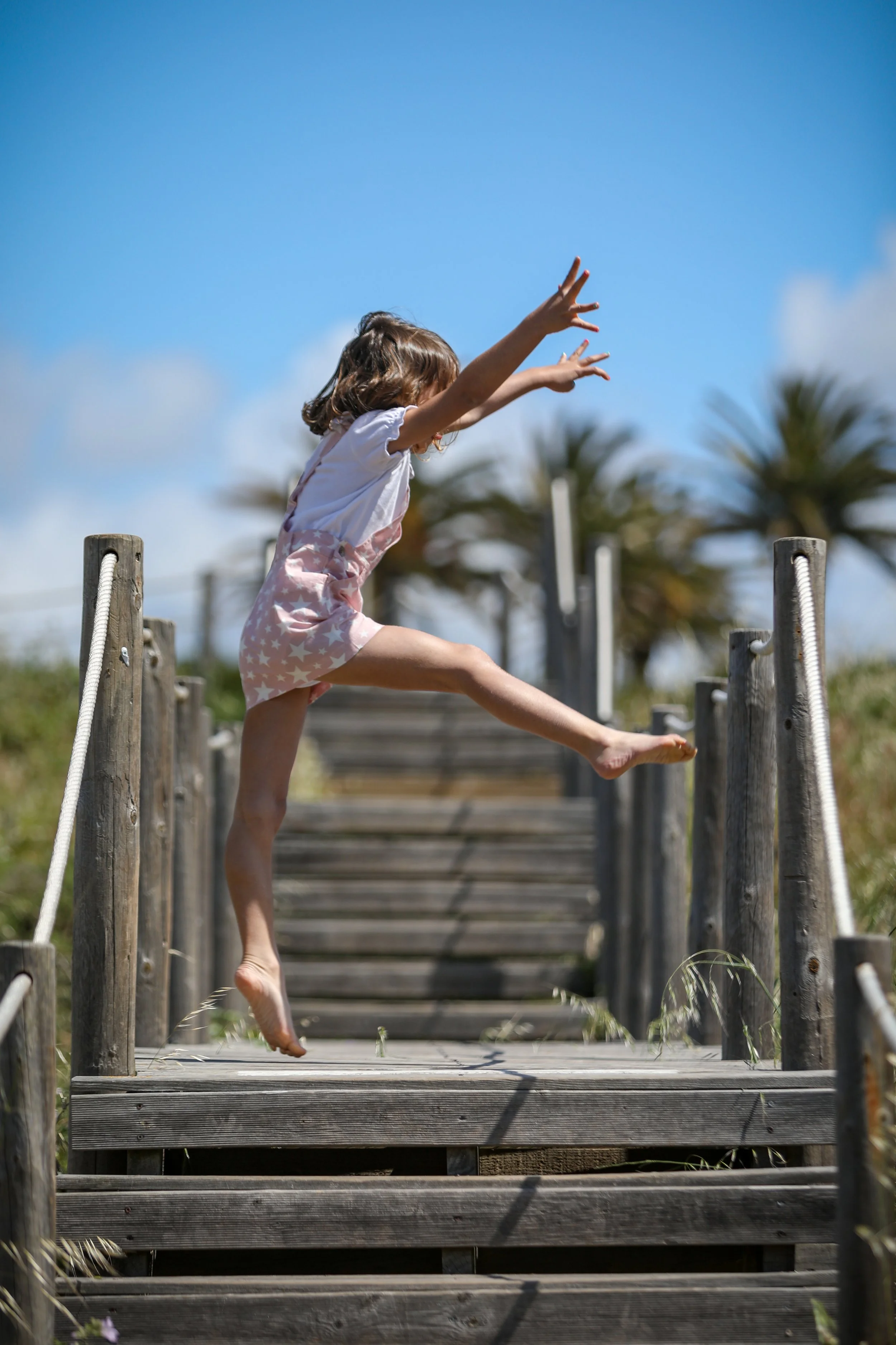 A young girl jumping off a wooden staircase on a sunny day with a bright blue sky and palm trees in the background.