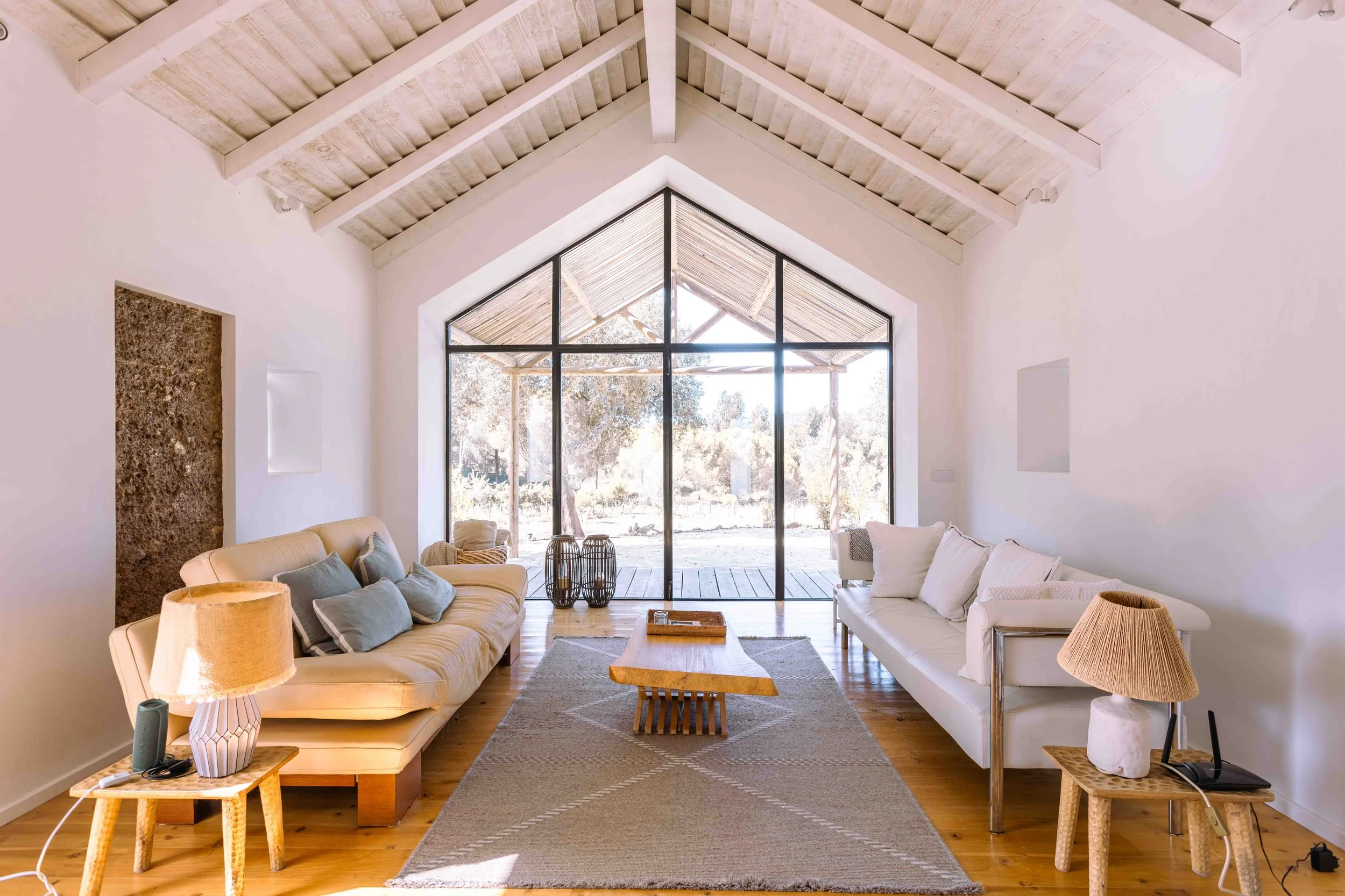 A living room with minimalist decor, featuring two cream-colored sofas with throw pillows, a wooden coffee table, a patterned rug, and side tables with lamps. Large glass doors open to a wooden patio with trees outside.