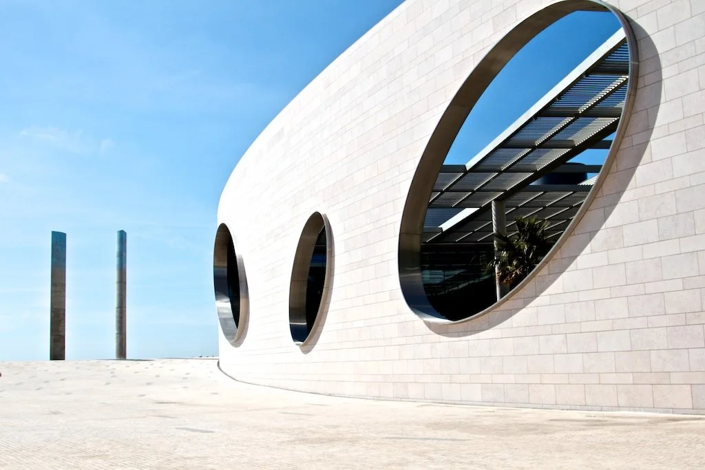 Modern white building with three large circular windows, blue sky, and three tall posts in the background.