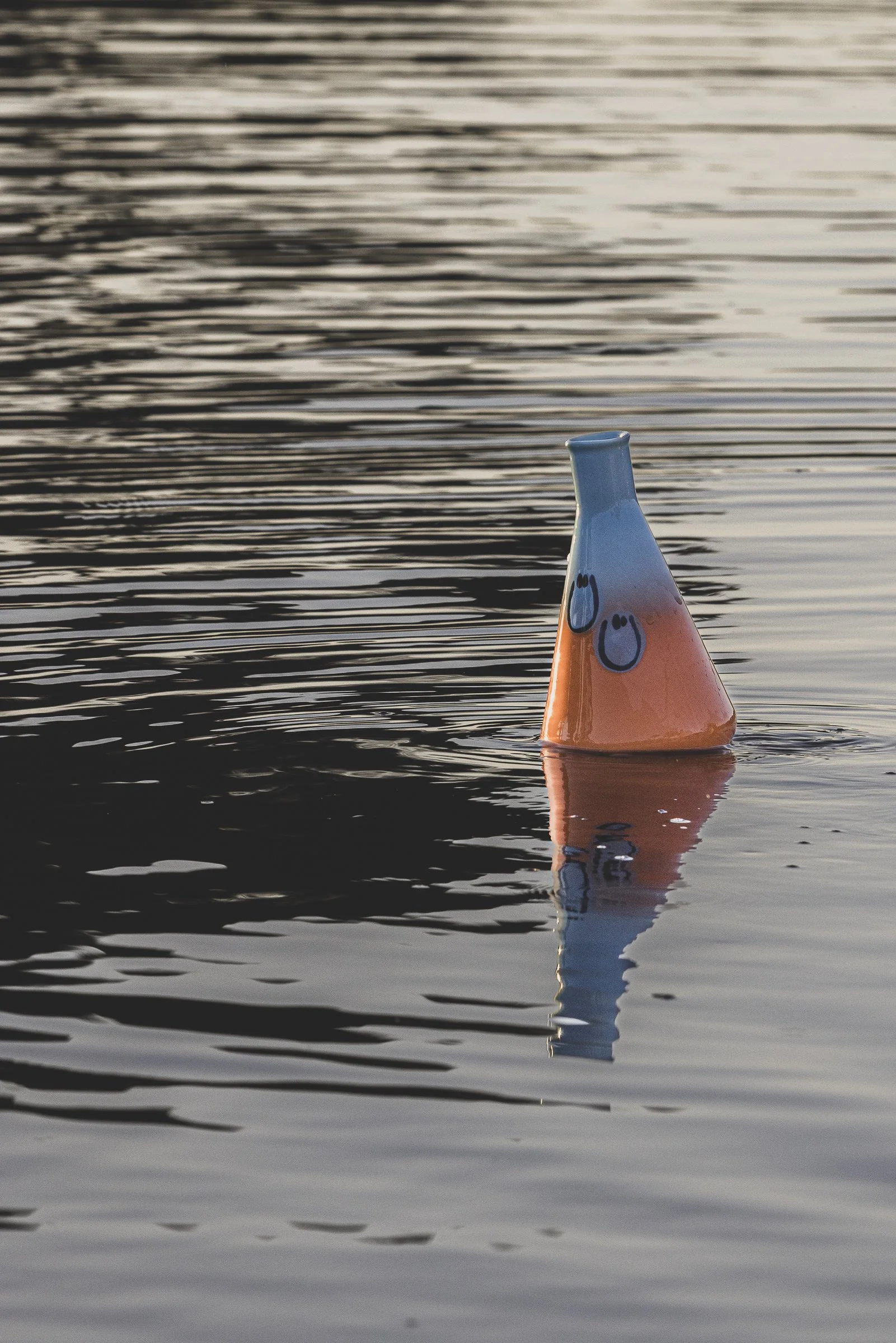 A colorful ceramic vase with a conical shape and abstract face design, floating upside down in calm water, with reflections and ripples.